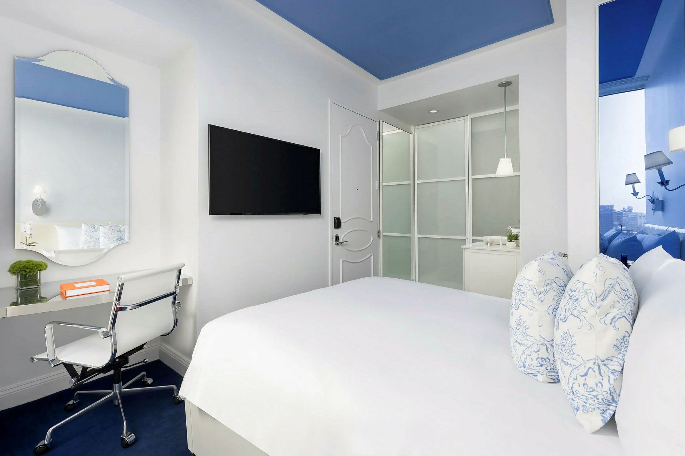 Ett modernt hotellrum med en dubbelsäng, skrivbord och stol samt en väggmonterad TV. Rummet är ljust med vita väggar och blå detaljer. (AI-genererad text)