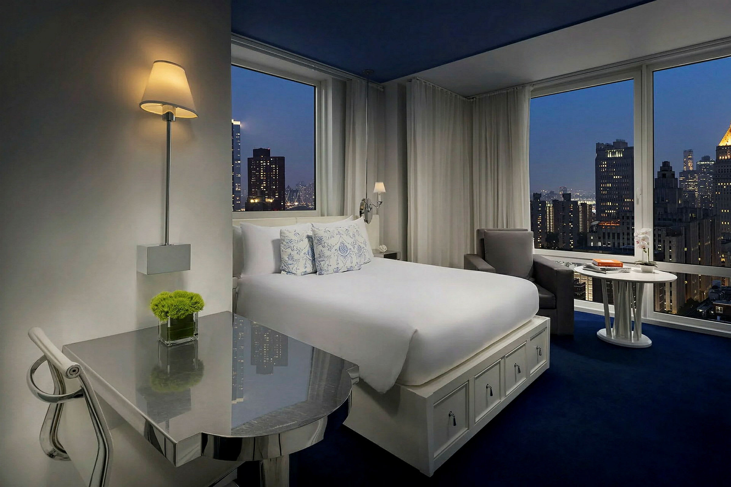 Hotellrum med stor säng och höga fönster med utsikt över stadens skyline. (AI-genererad text)