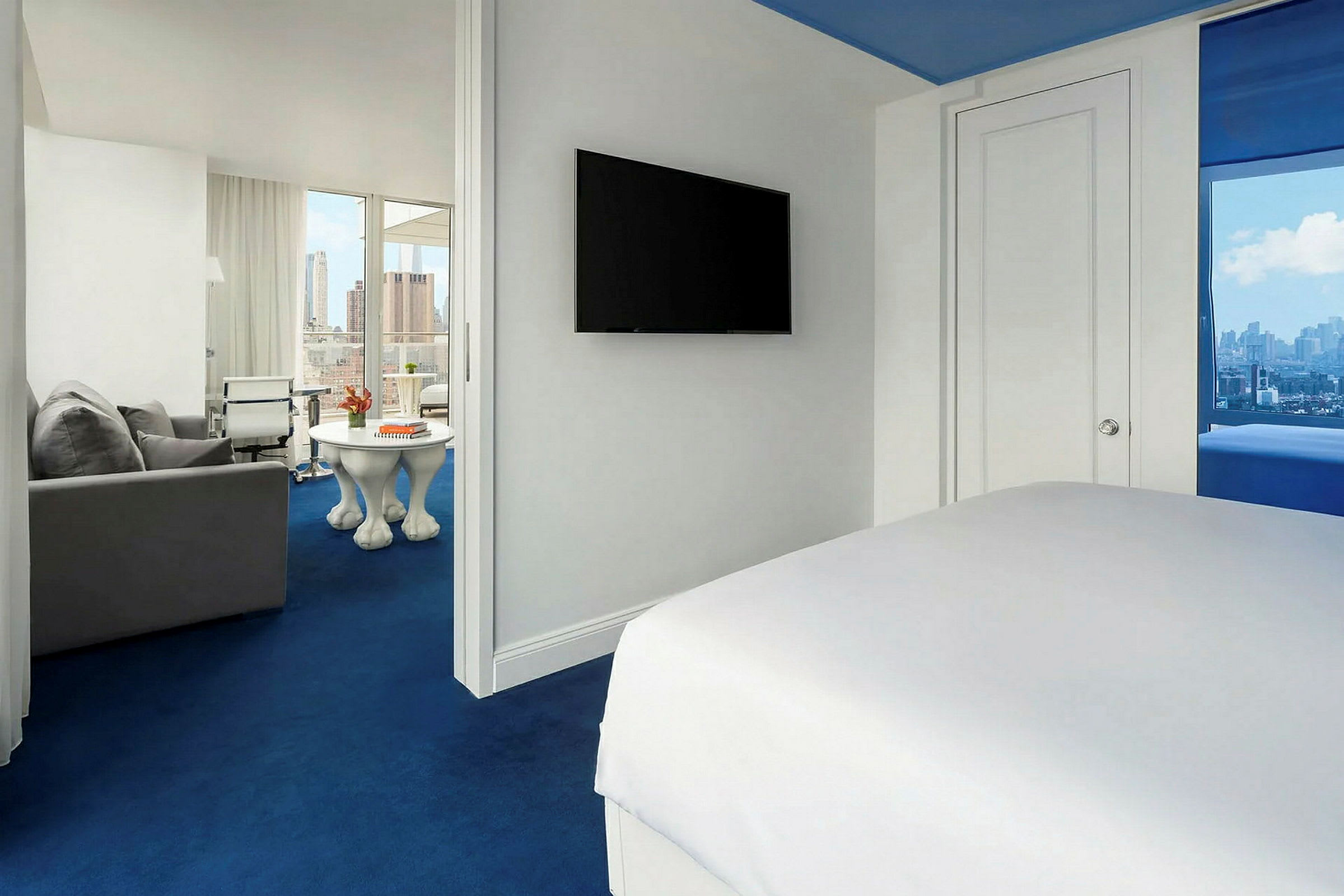 Hotellrum med vit inredning och blå matta. Sängen syns delvis i förgrunden, en TV hänger på väggen och i bakgrunden finns en sittgrupp med utsikt över staden. (AI-genererad text)