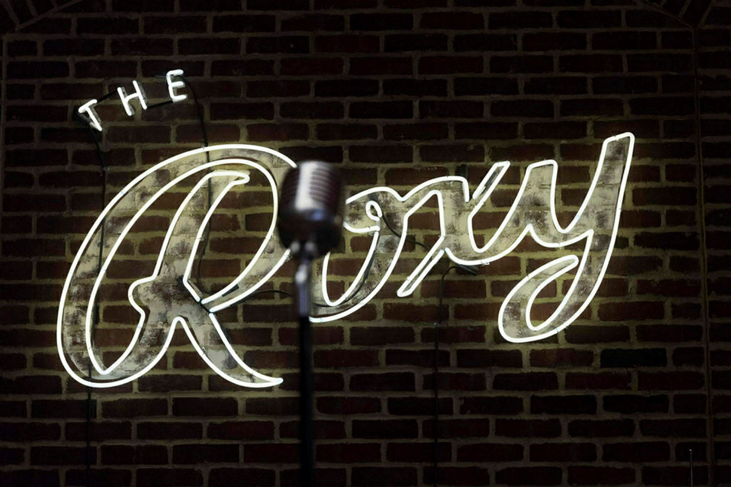 En mikrofon framför en neonskylt med texten 'The Roxy' på en tegelvägg. (AI-genererad text)