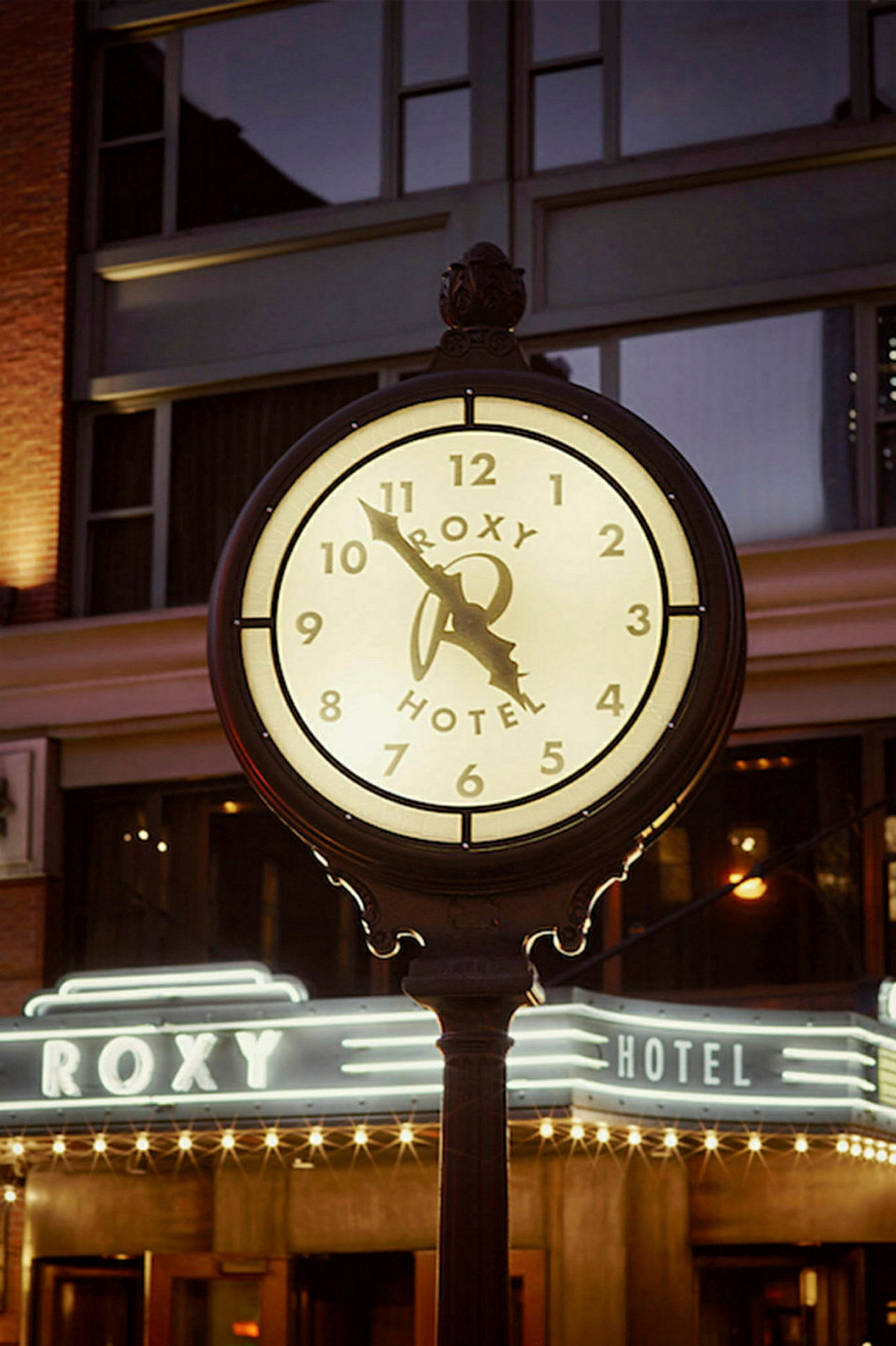En upplyst klocka med texten "Roxy Hotel" framför en byggnad på kvällen. (AI-genererad text)