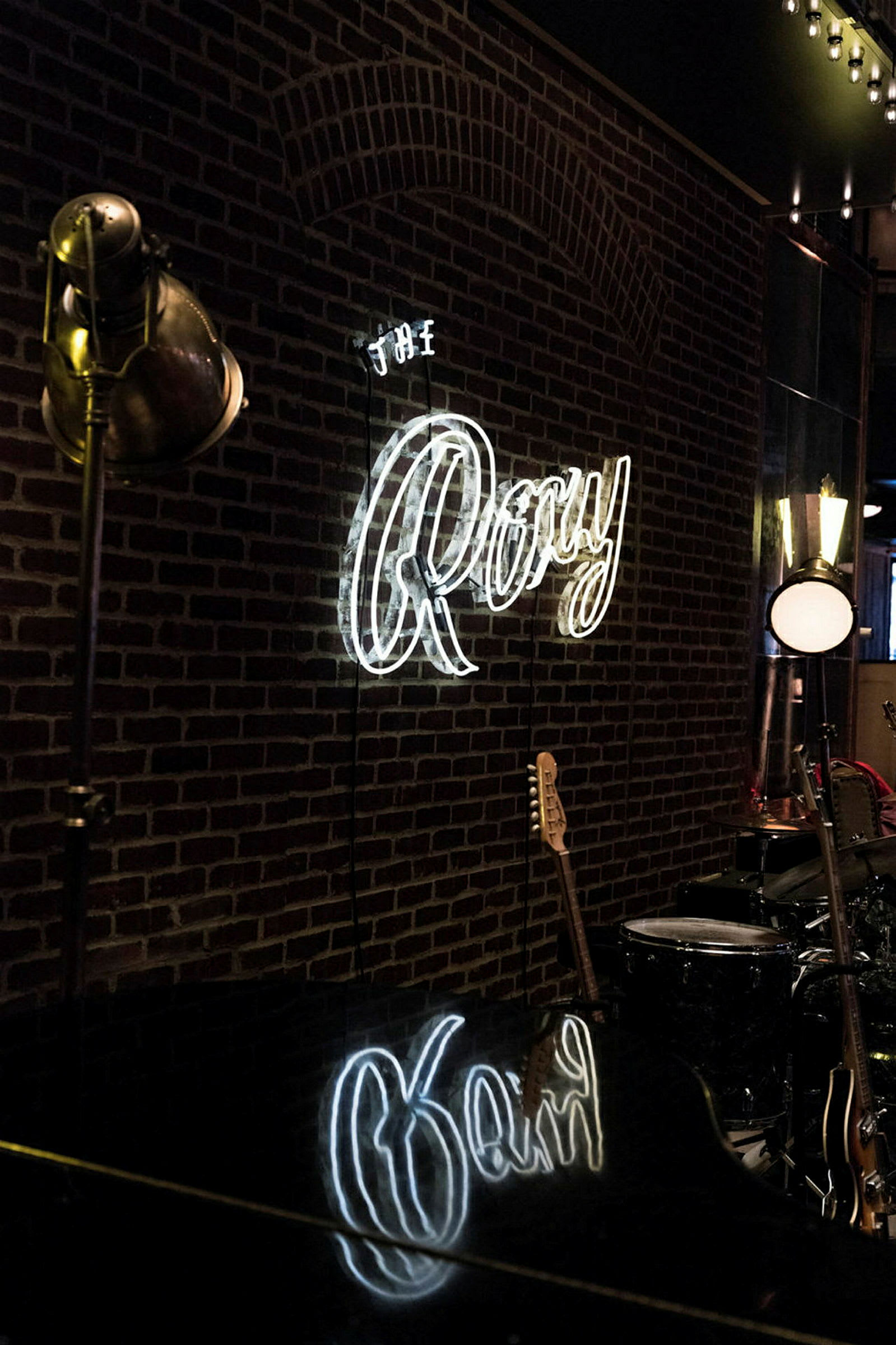 En vägg med en neonlampa som skriver "The Roxy" i vita bokstäver, samt musikinstrument som en gitarr och trummor synliga i bakgrunden. (AI-genererad text)