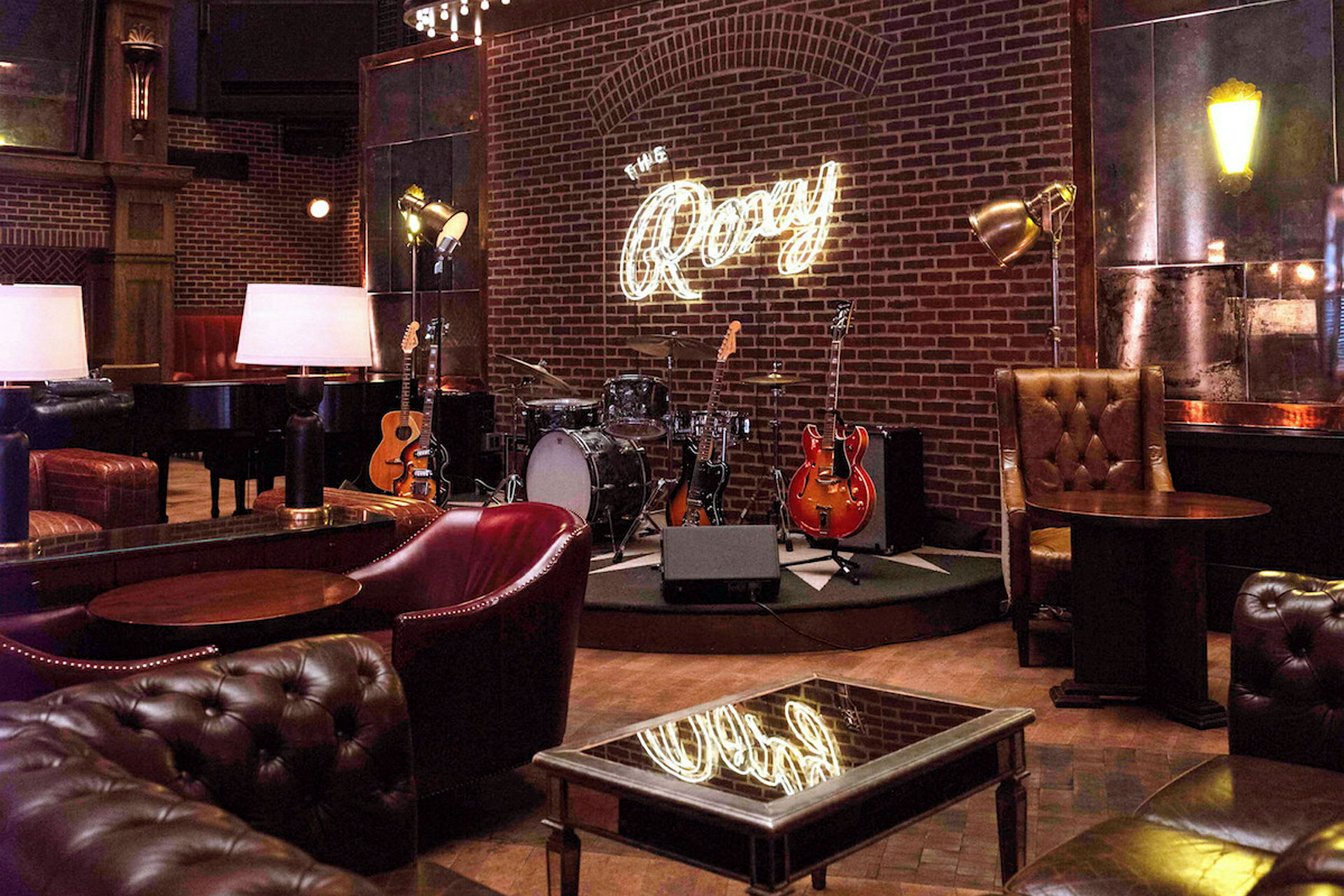 Interiör av en musikklubb med gitarrer, ett trumset och en liten scen under en neonskylt som lyder 'The Roxy'. (AI-genererad text)