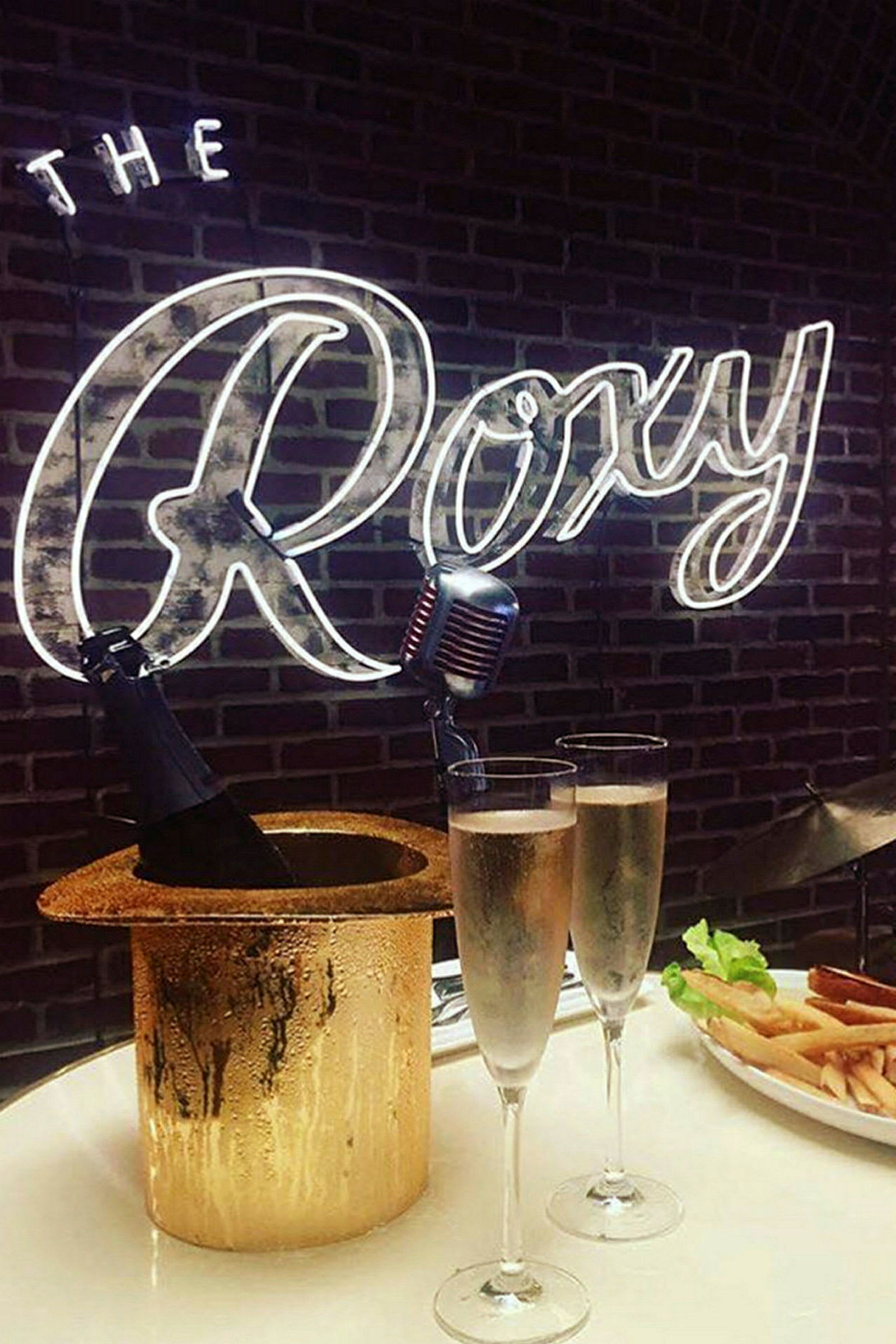 Ett bord med två glas mousserande dryck, champagneflaska i en ishink formad som en hatt, samt en tallrik med pommes frites. Neontexten "The Roxy" syns i bakgrunden. (AI-genererad text)