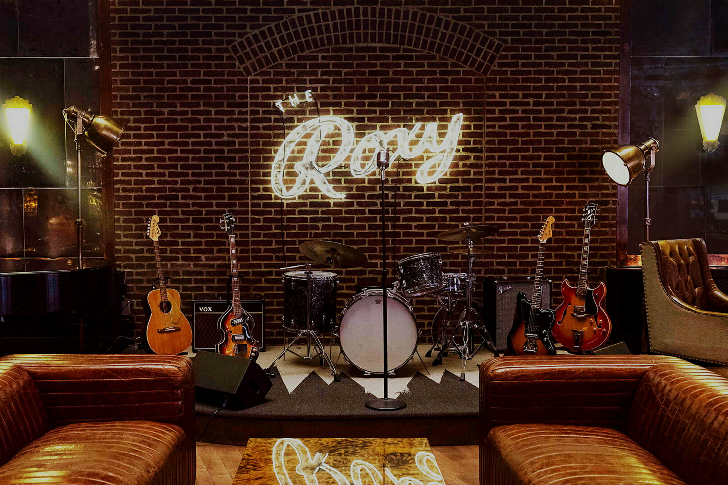 En scen med musikinstrument som trummor och gitarrer framför en tegelvägg med en lysande skylt som säger "The Roxy". (AI-genererad text)
