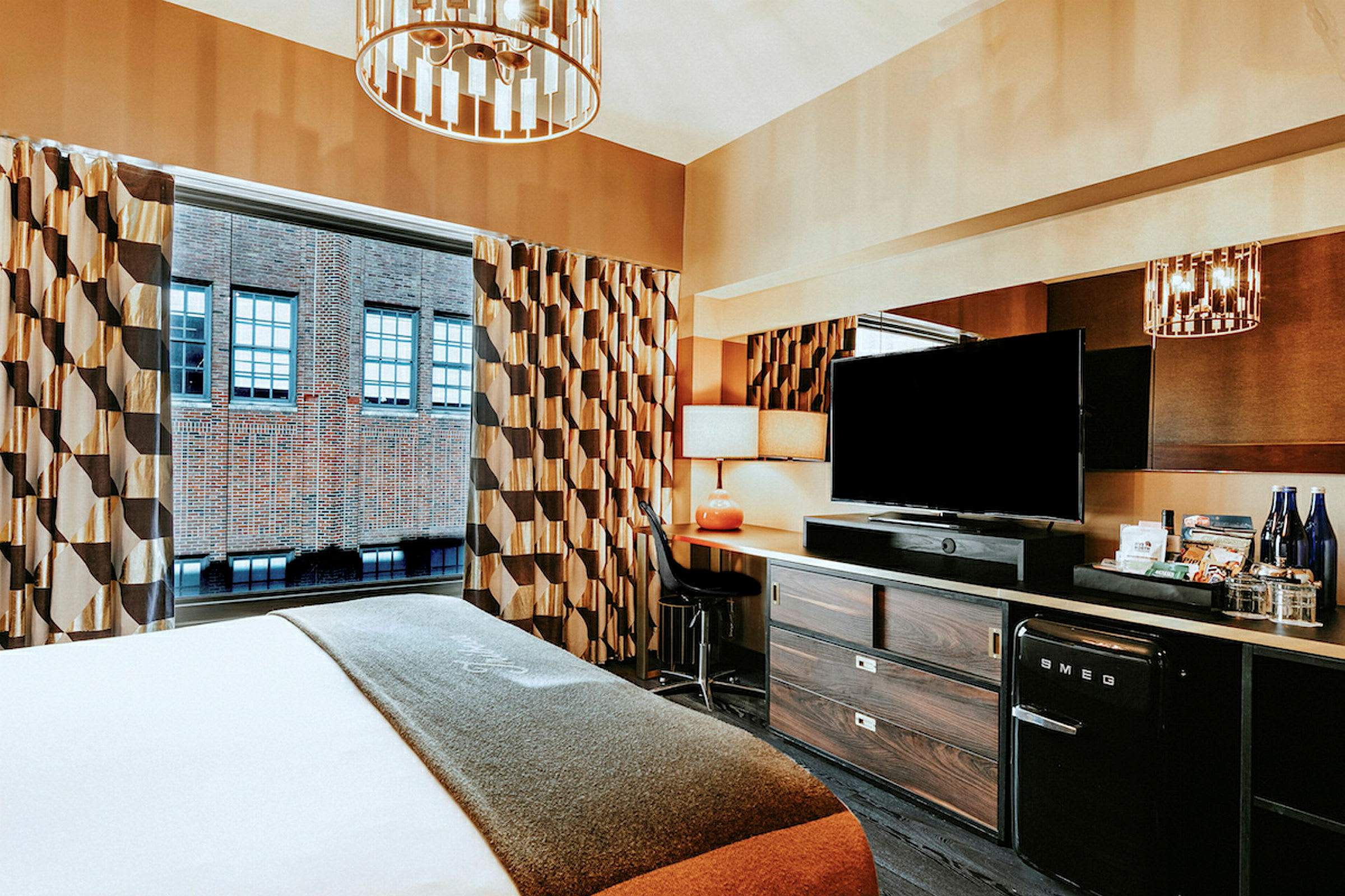 Hotellrum med en säng, skrivbord och TV, samt fönster mot en tegelbyggnad. (AI-genererad text)