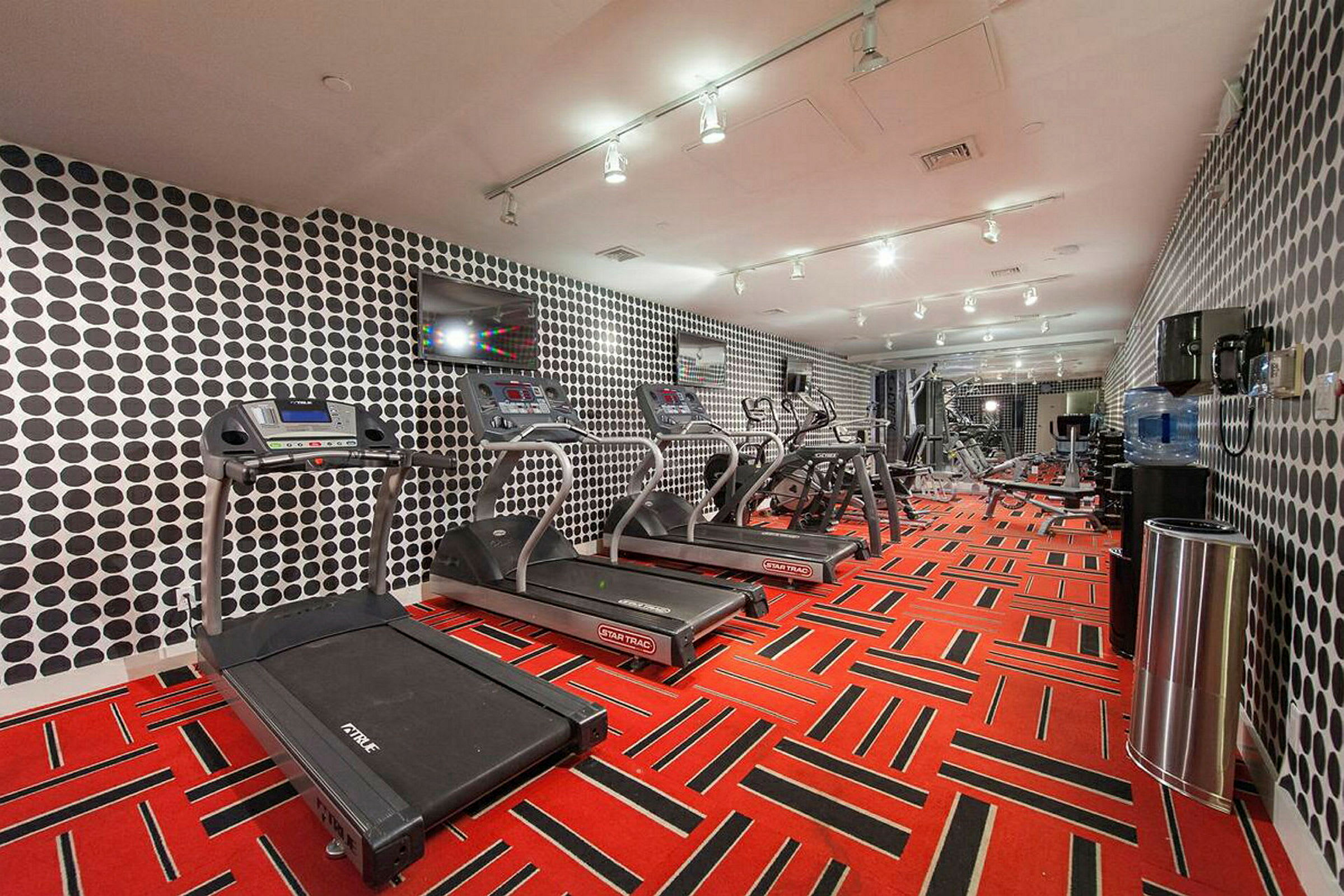 Ett gym med löpband, motionscyklar och andra träningsmaskiner. Väggarna har ett cirkulärt mönster, och golvet är rött med svarta detaljer. (AI-genererad text)