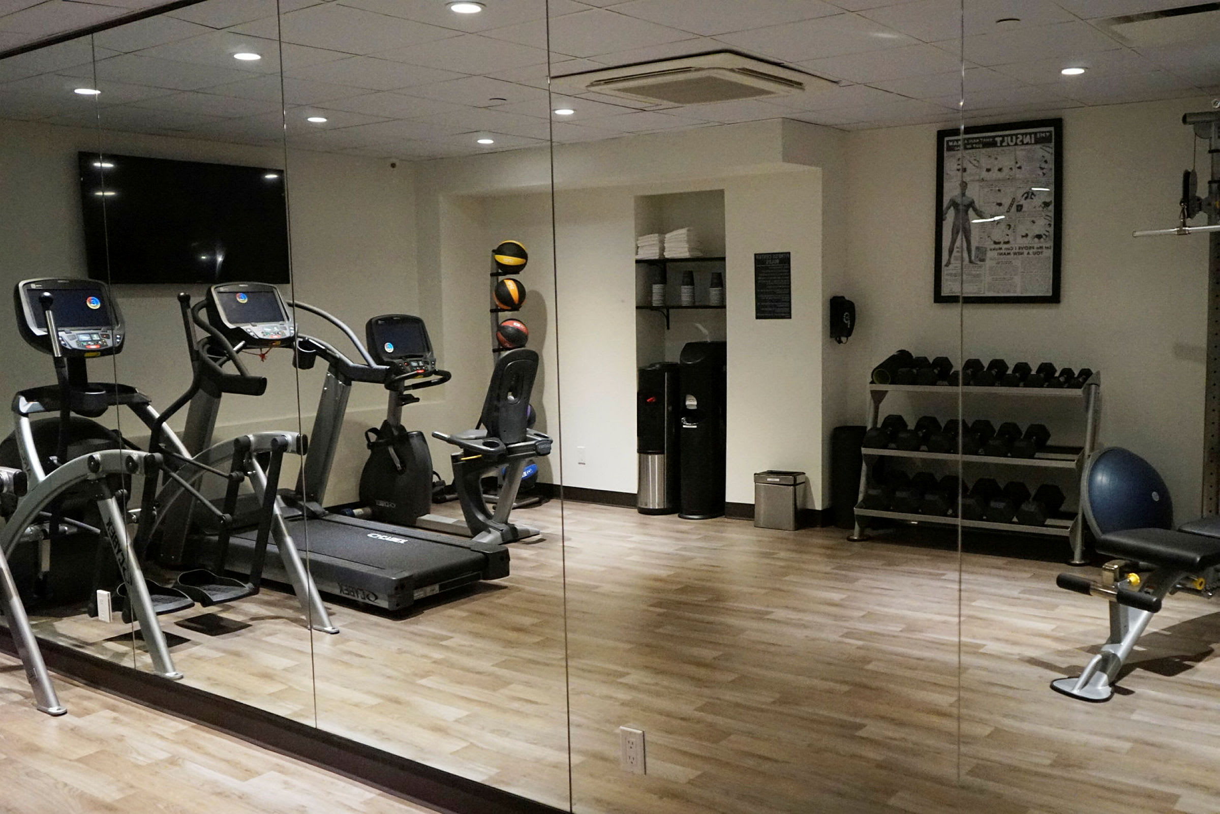 Ett gym med spegelvägg, träningsmaskiner, hantlar och annan träningsutrustning syns. (AI-genererad text)
