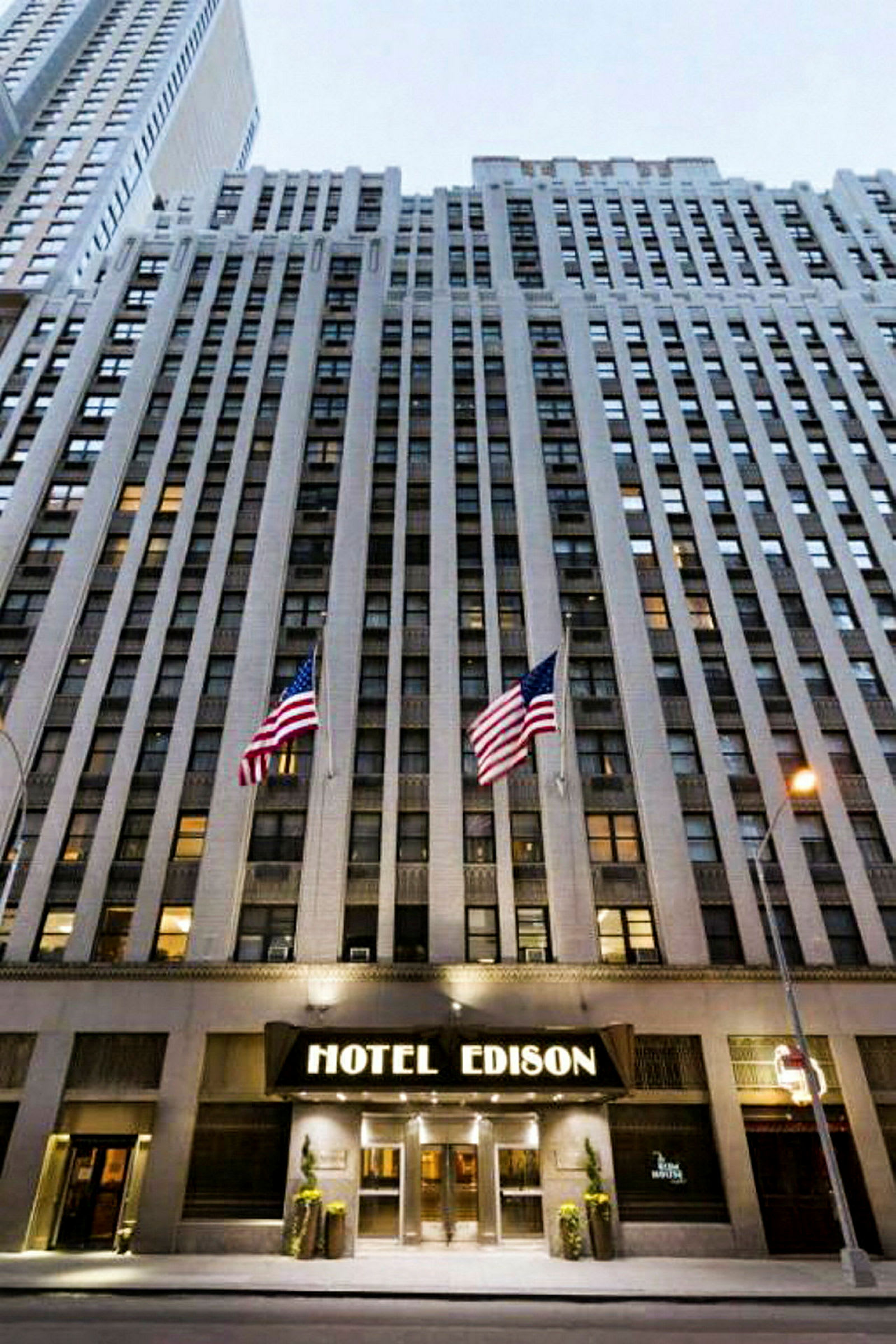 Utsikt mot en hög byggnad med ingången märkt "Hotel Edison" och två amerikanska flaggor framför. (AI-genererad text)