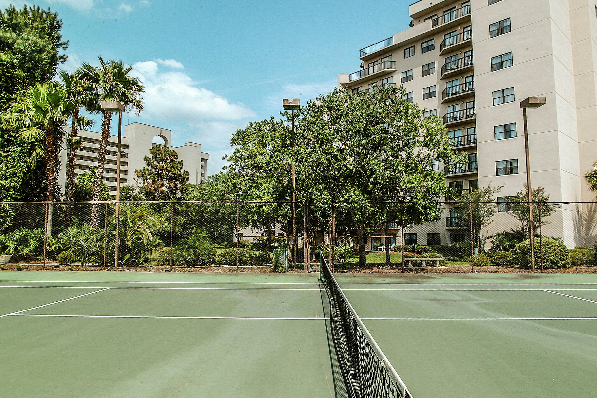 Tennisbana omgiven av gröna växter och byggnader i bakgrunden. (AI-genererad text)