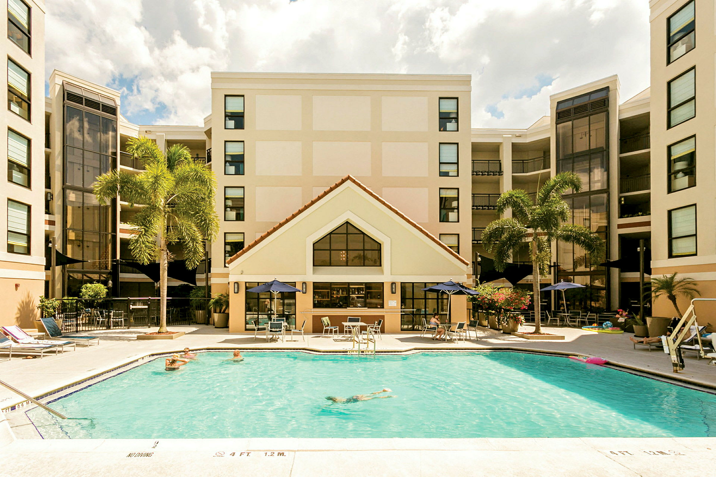 Bild av ett hotell med en pool i förgrunden. Personer badar och solar vid poolområdet. Hotellets fasad syns i bakgrunden med palmer i närheten. (AI-genererad text)