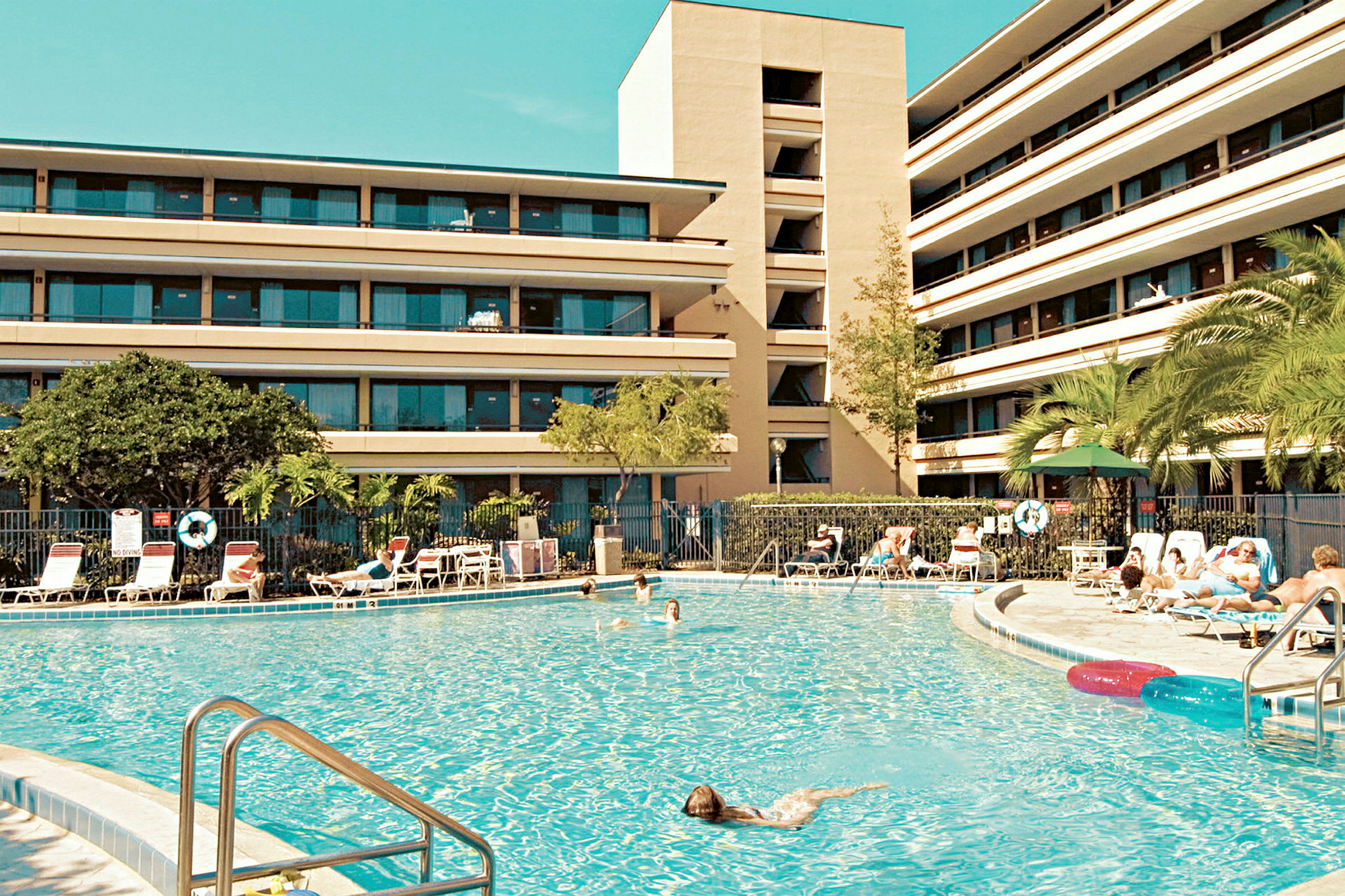 En stor swimmingpool omgiven av solstolar och en fler vånings byggnad i bakgrunden. (AI-genererad text)