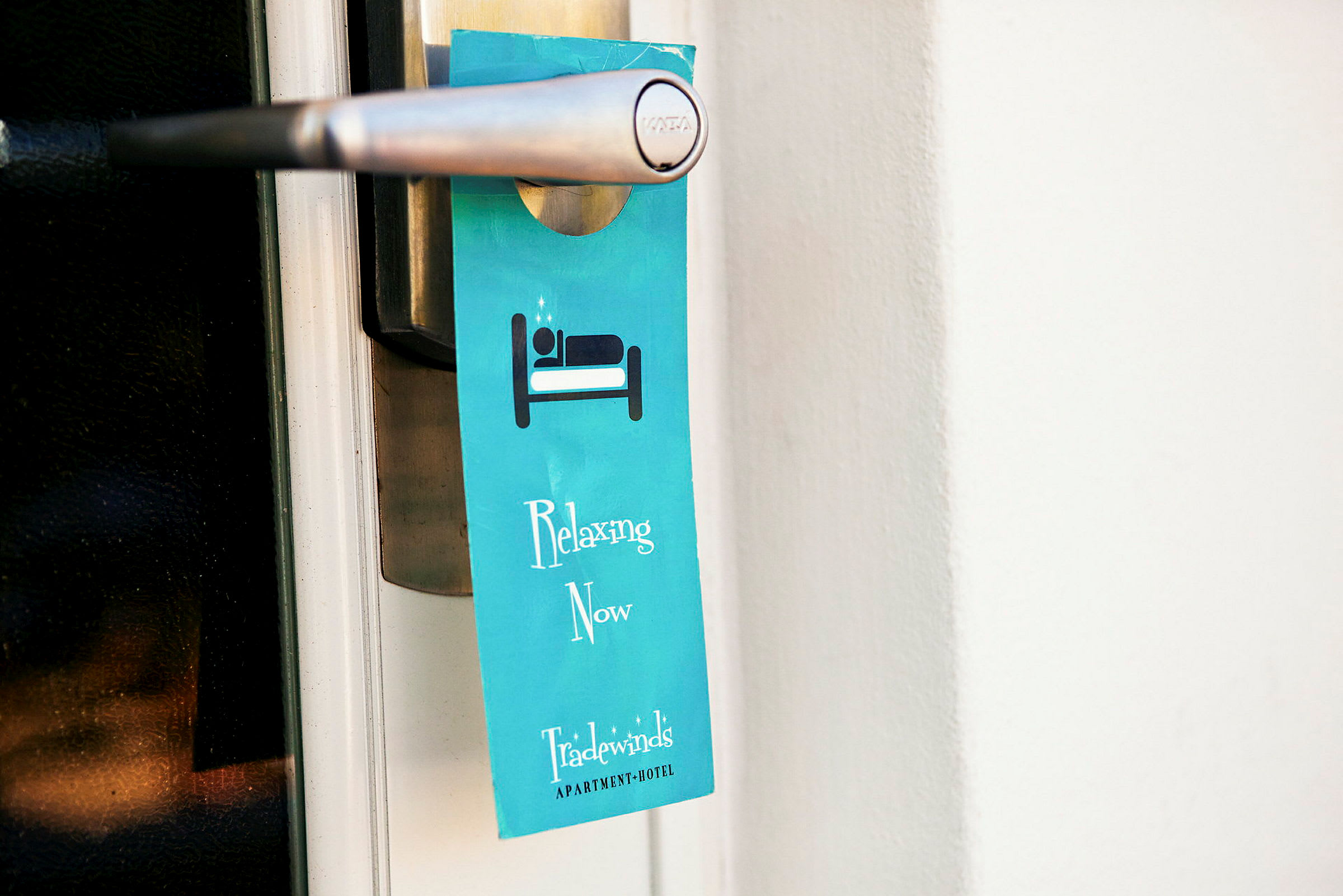 En blå skylt hänger på ett dörrhandtag och visar texten "Relaxing Now" samt är märkta med hotellets namn. (AI-genererad text)