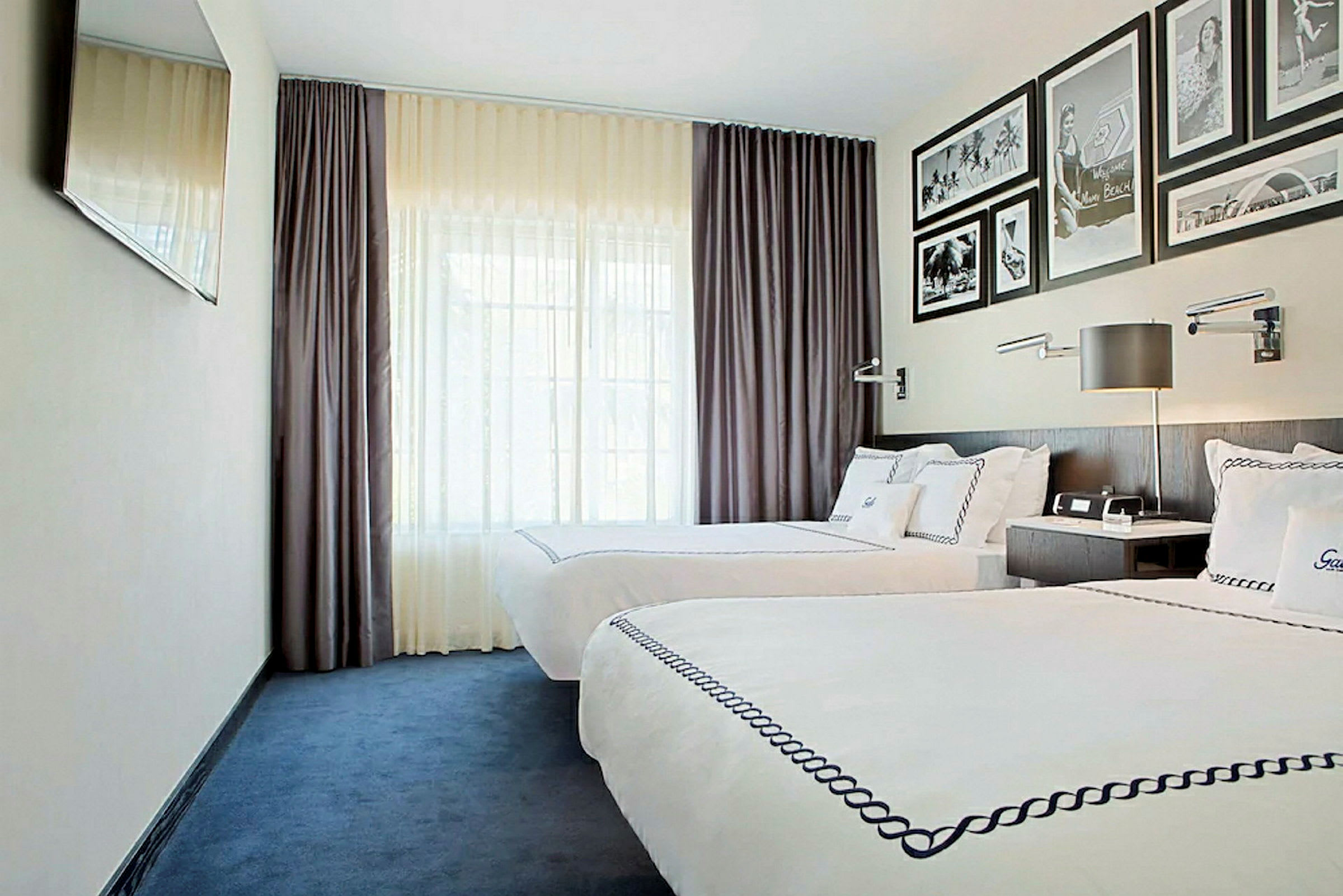 Ett hotellrum med två enkelsängar med vita lakan och dekorerade överkast. Rummet har mörklila gardiner, en väggmonterad TV och inramade bilder på väggen över sänggaveln. (AI-genererad text)
