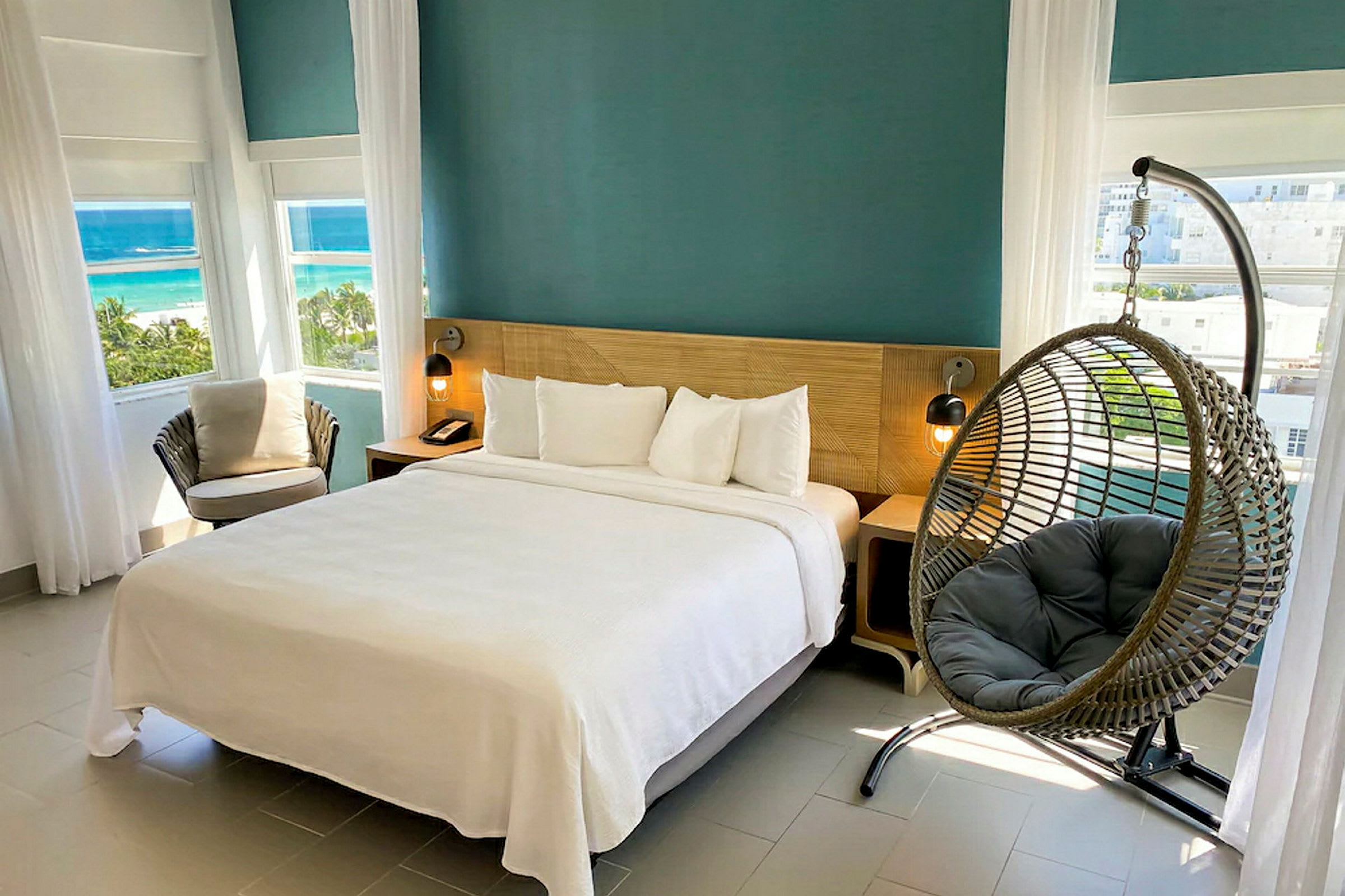 Ett hotellrum med en dubbelsäng, en hängstol och stora fönster med utsikt över stranden. (AI-genererad text)