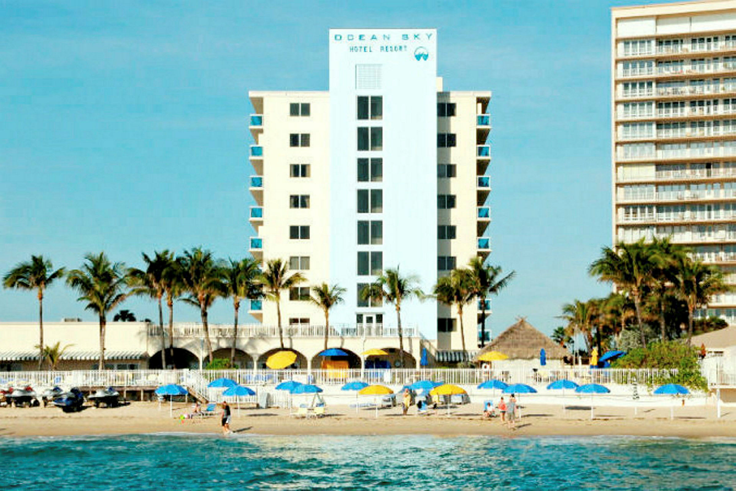 Ett hotell med palmträd framför, bredvid stranden med parasoller och människor på sanden. (AI-genererad text)