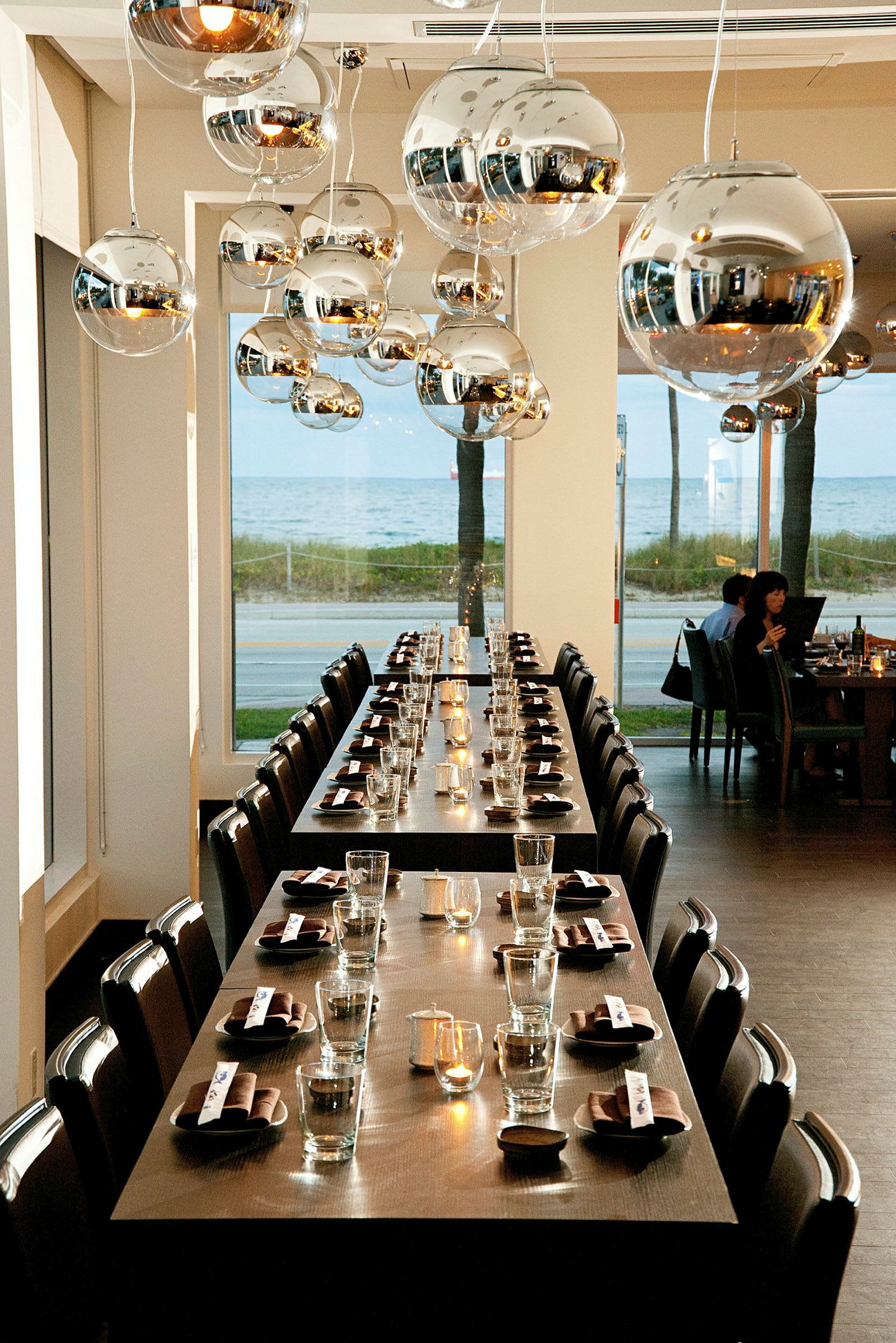 Bilden visar en restaurang med långbord som är dukade med glas och servetter. Taket har moderna metalliska ljuskronor, och det finns höga fönster med utsikt över havet. (AI-genererad text)