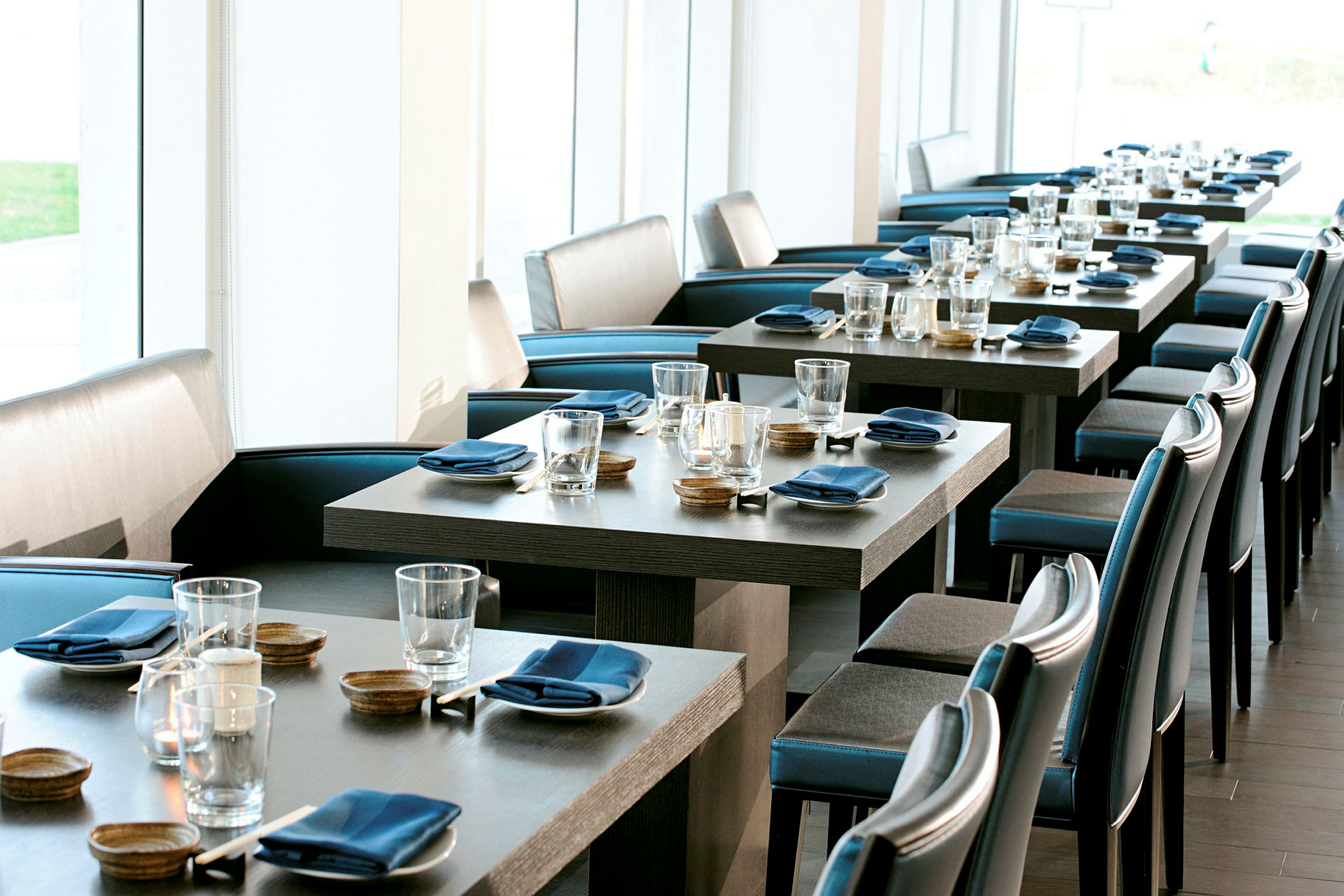 Ett elegant restaurangområde med dukade bord, blå servetter och glas. Ljusa fönster i bakgrunden. (AI-genererad text)