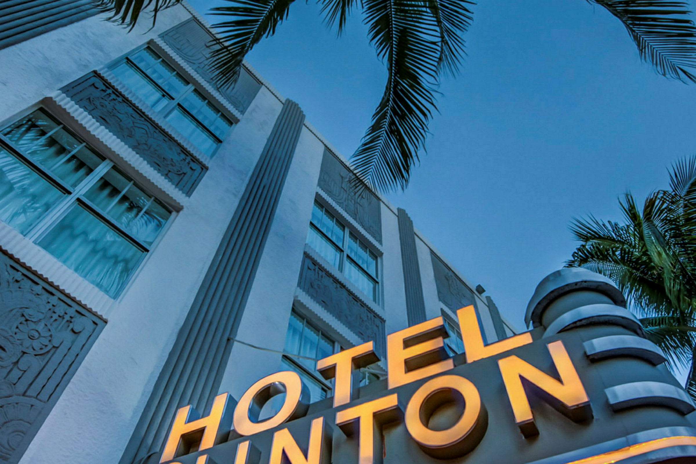 En närbild av en vit byggnad med en upplyst skylt som lyder "HOTEL" och palmer i förgrunden mot en klarblå himmel. (AI-genererad text)
