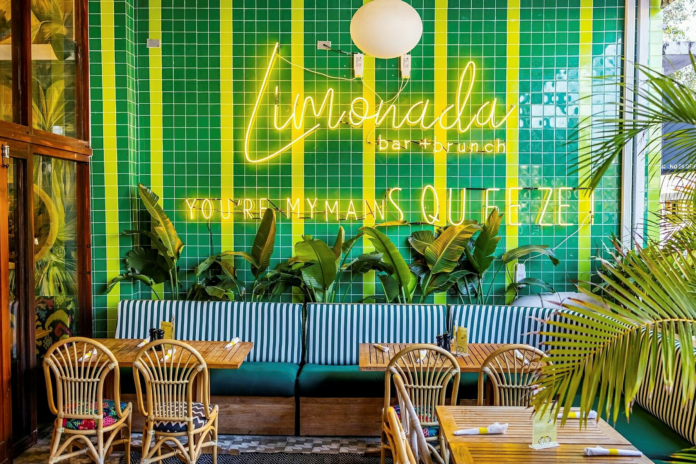 Restauranginteriör med grönt kaklat väggytor, neonskyltar, palmer, träbord och flätade stolar. (AI-genererad text)