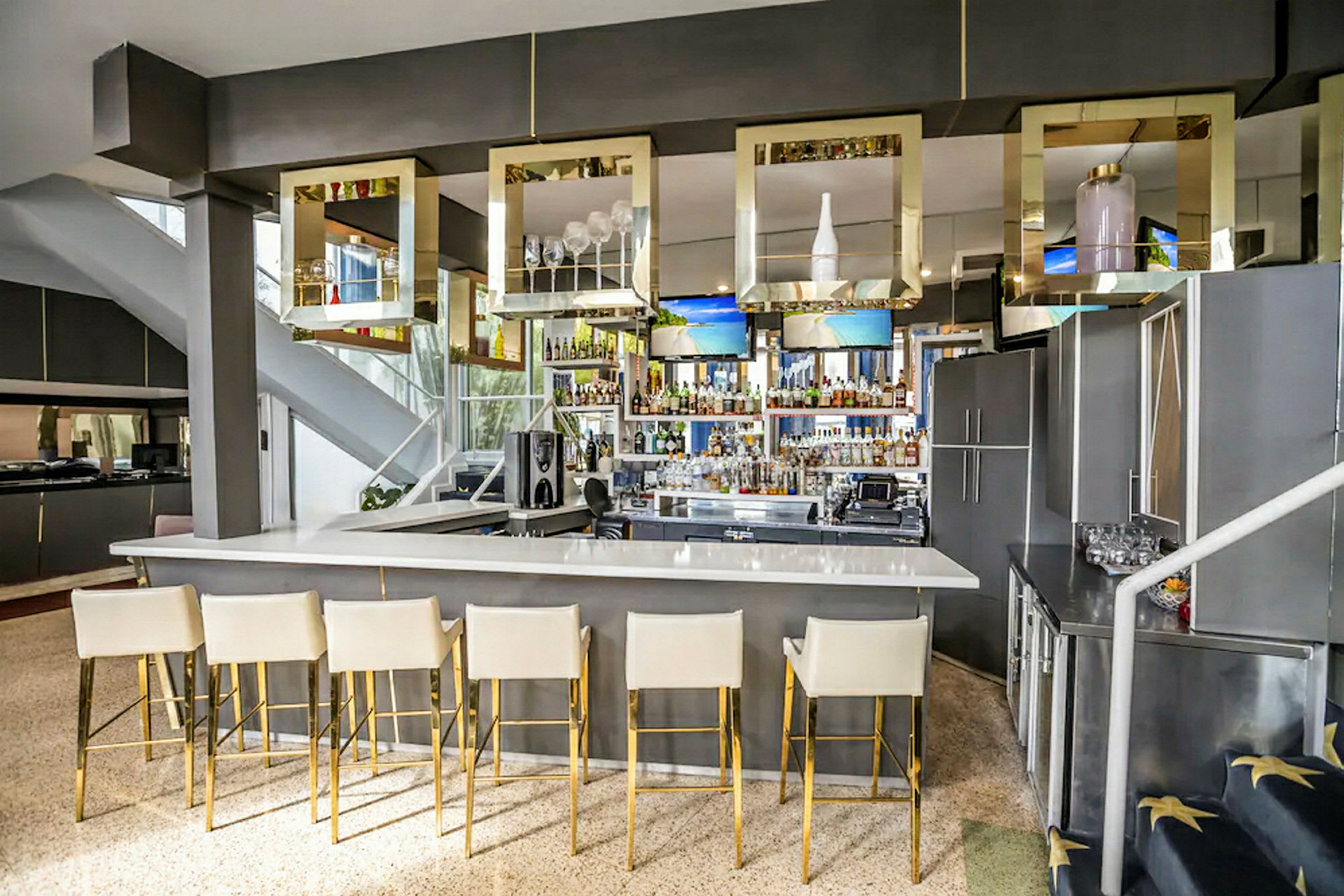 En modern bar med vit bardisk och högryggade stolar i ljus färg och gyllene ben. Över bardisken hänger fyrkantiga dekorationer. (AI-genererad text)
