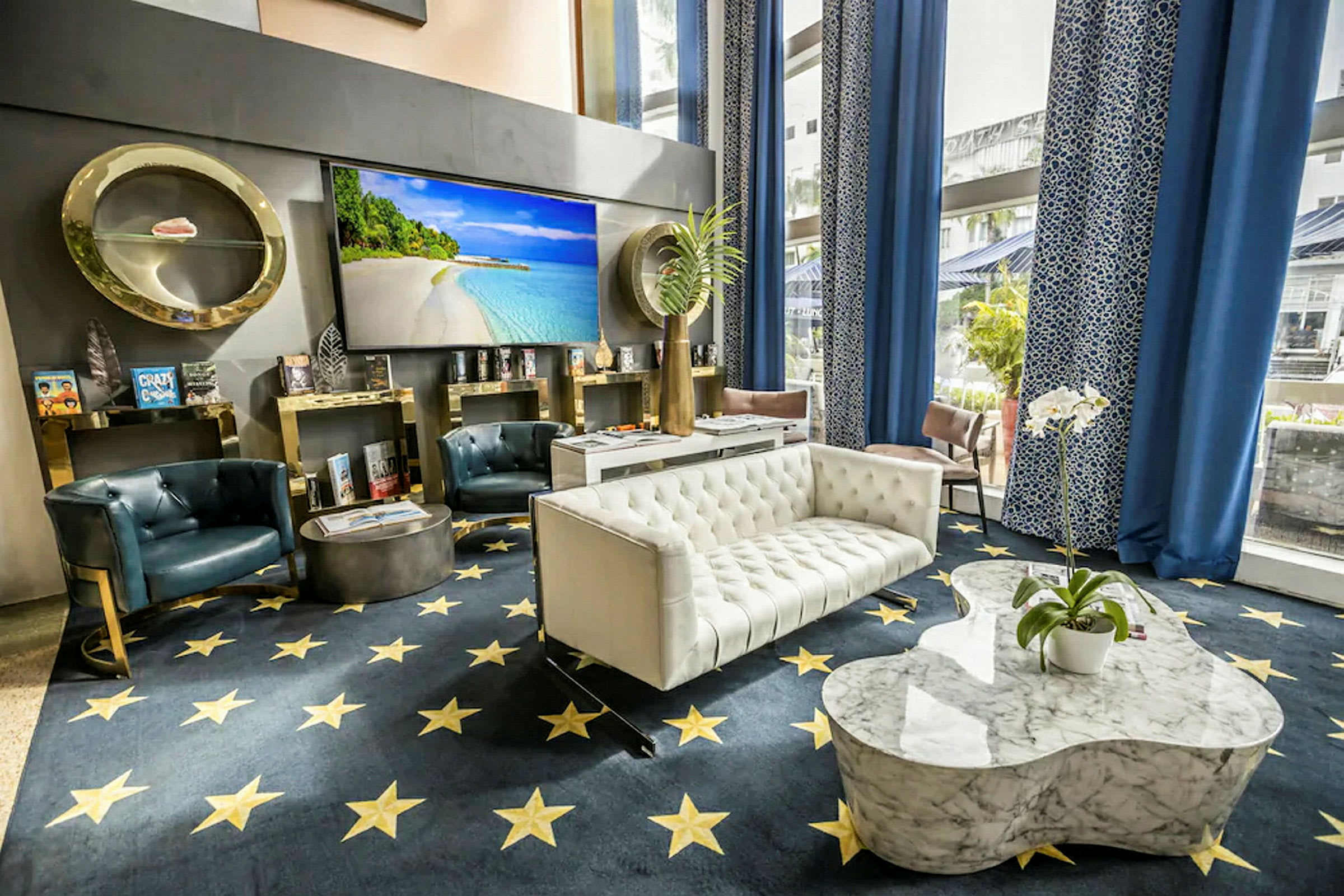 Bild av en elegant hotellobby med soffor, fåtöljer och ett marmorbord. Bakgrunden har en stor väggskärm med strandmotiv och inredningen inkluderar blå draperier och dekorativa detaljer. (AI-genererad text)