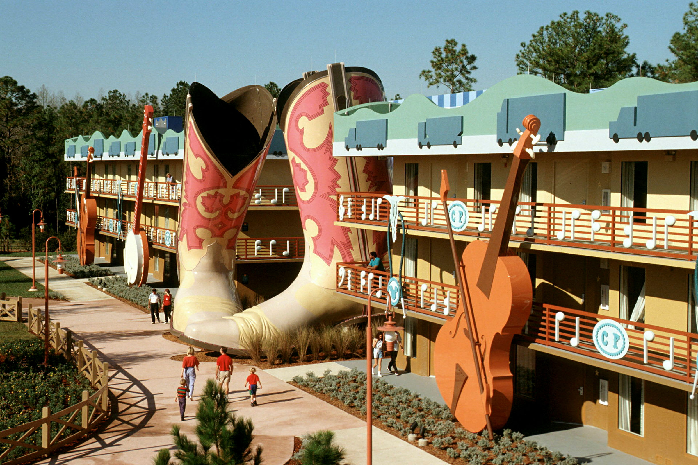 Ett färgglatt hotellkomplex med stora cowboyboots och en gitarr i dekor. Folk promenerar på gångvägen. (AI-genererad text)