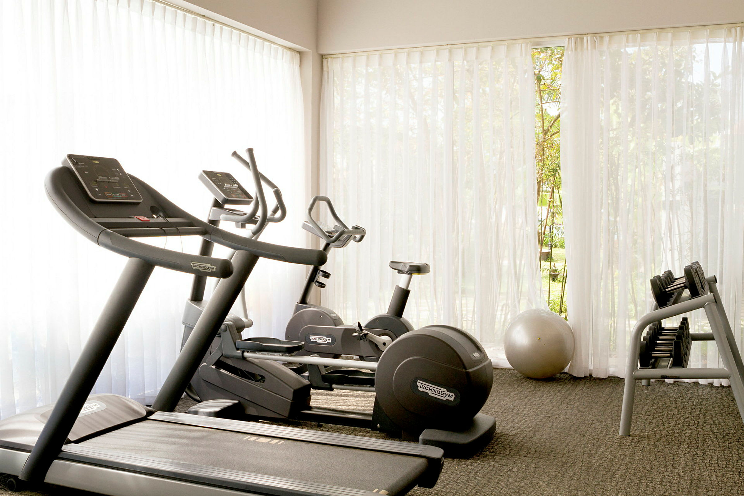Ett gym med träningsmaskiner inklusive löpband, motionscykel och hantlar, med naturligt ljus som strömmar genom stora fönster. Vita gardiner skapar en ljus och luftig atmosfär. (AI-genererad text)