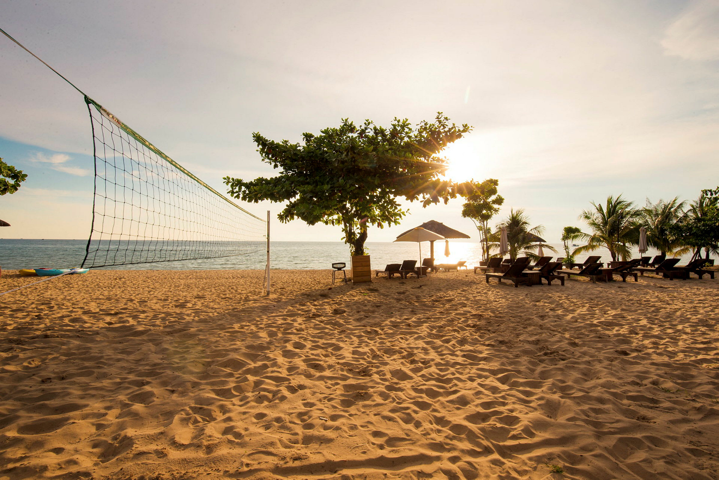 En sandstrand med en volleybollnät, solstolar under parasoll och några träd. Havet syns i bakgrunden under en solnedgång. (AI-genererad text)