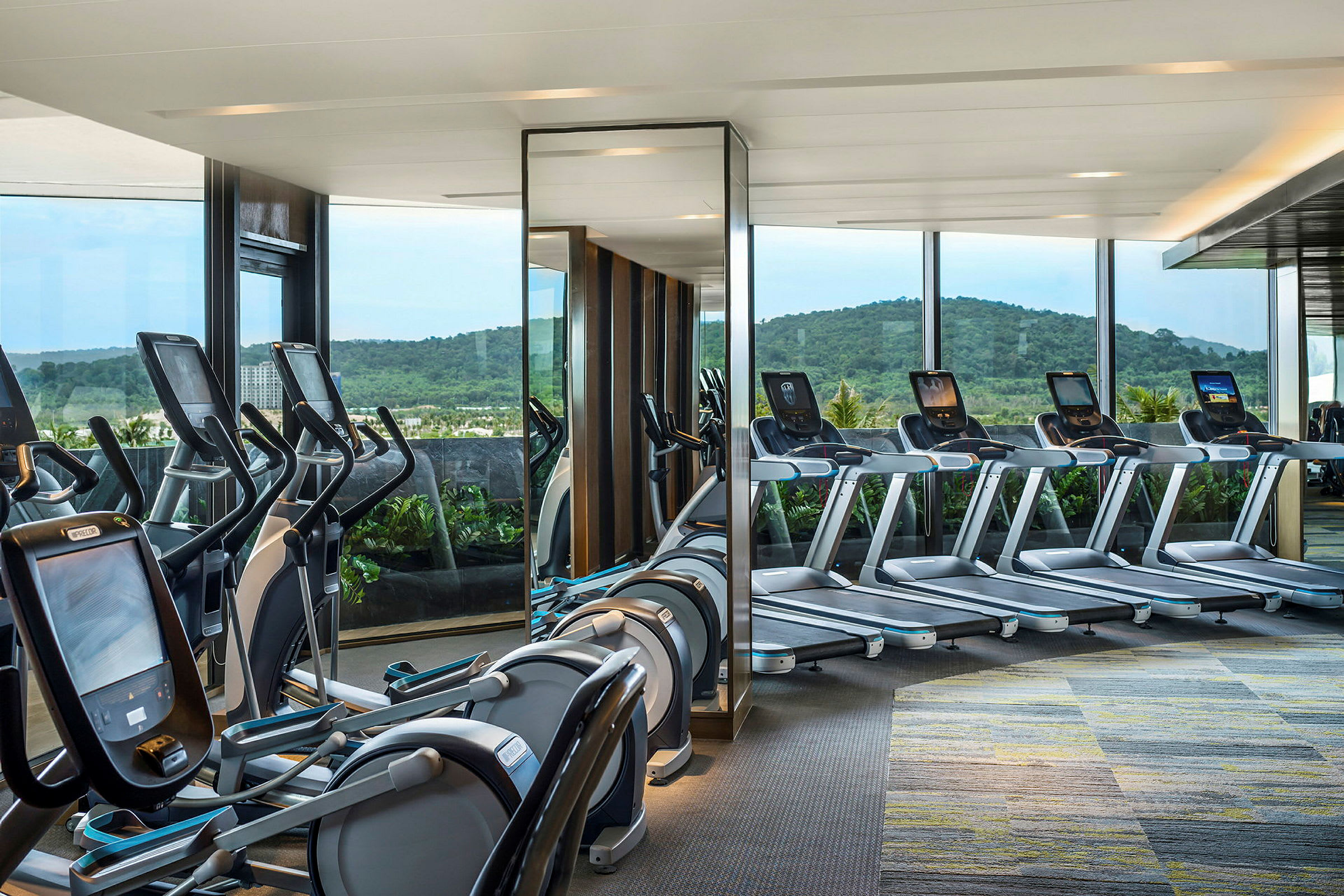 Ett gym med löpband och träningsmaskiner placerade framför stora fönster med utsikt över en grönskande bergsmiljö. (AI-genererad text)