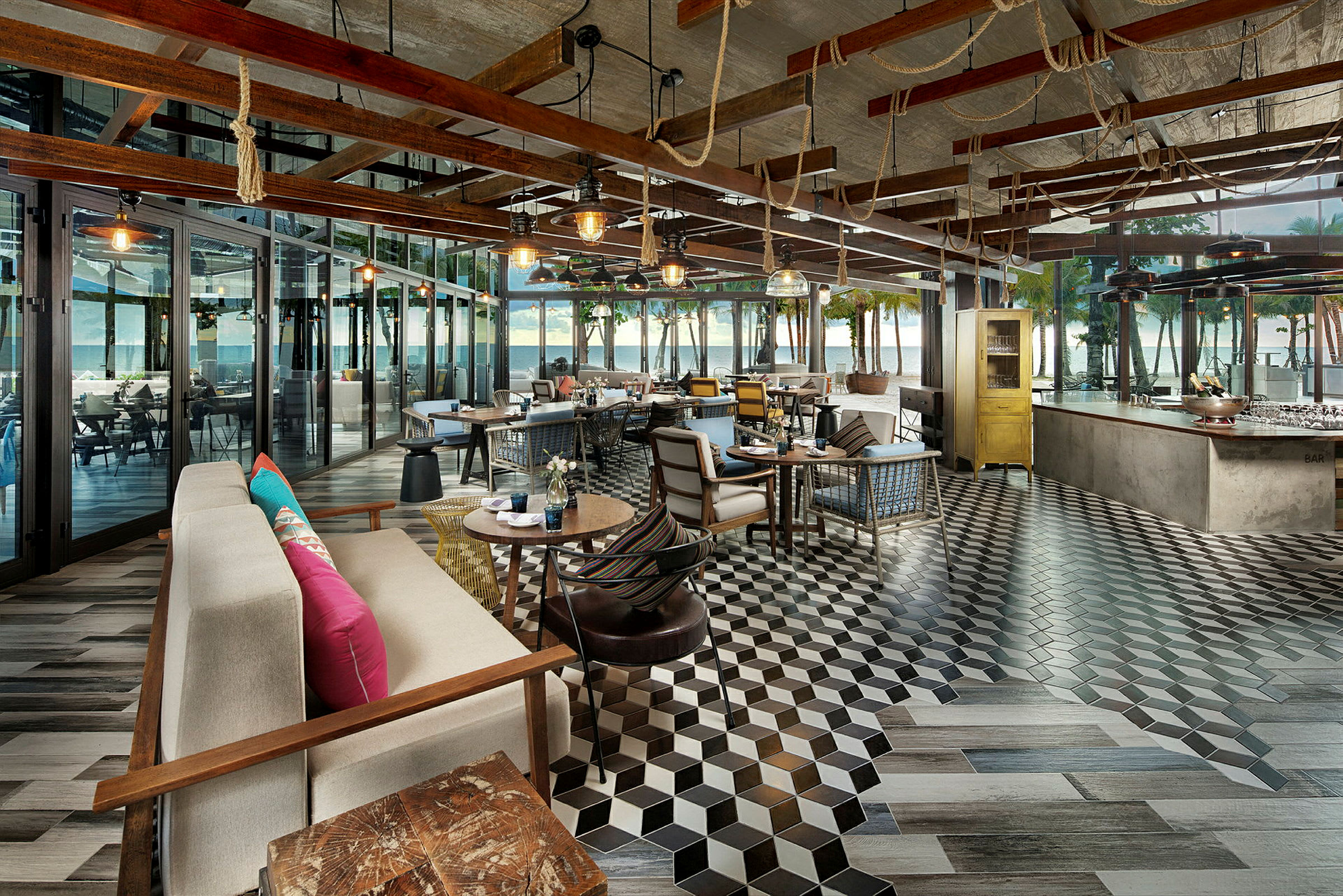 Ett café eller restaurang med modern inredning, sittgrupper och bord, samt stora glaspartier som vetter mot en terrass med utsikt över havet och palmer. (AI-genererad text)