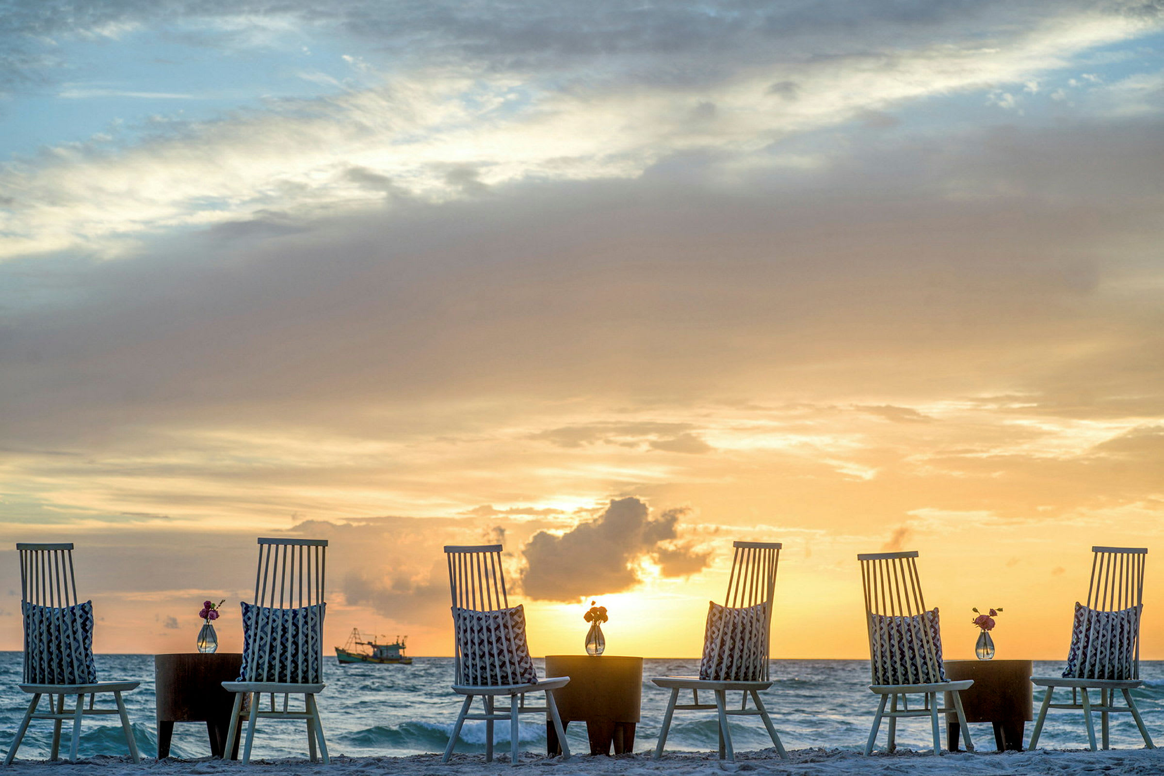 Stolar och bord på en strand med havet och solnedgång i bakgrunden. (AI-genererad text)
