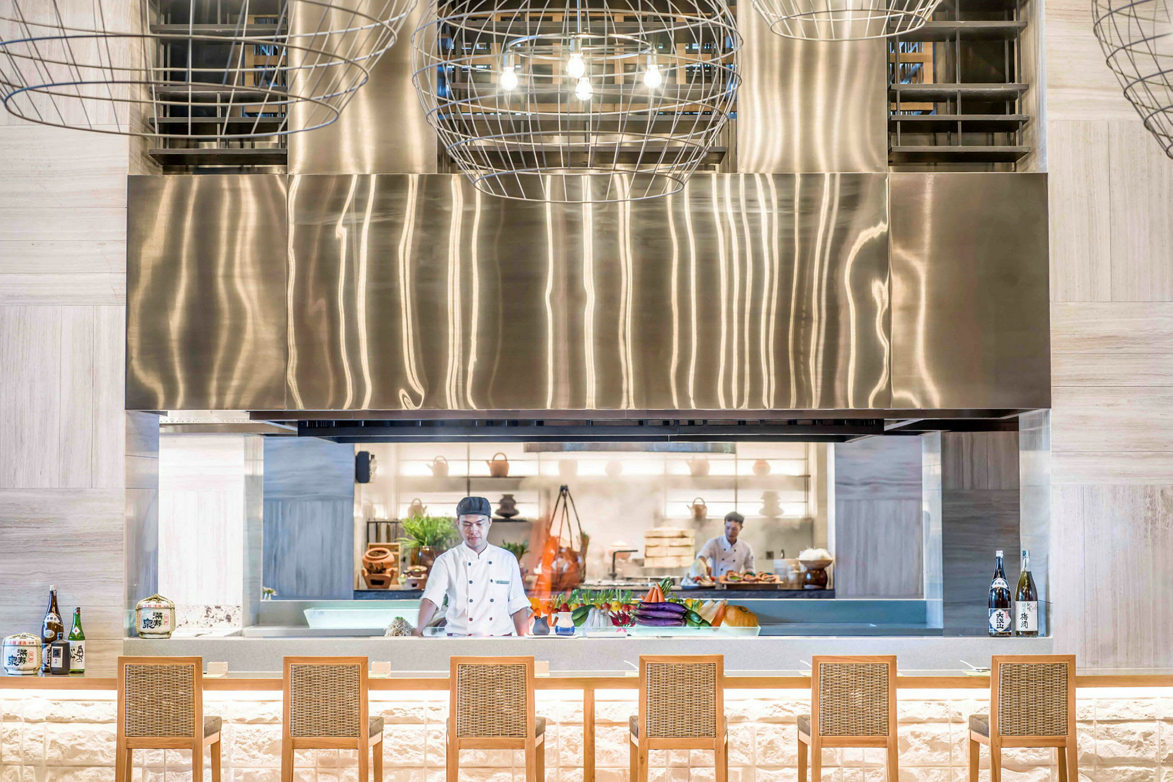En modern restaurang med en öppen köksmiljö. En kock arbetar bakom en stor, utställningsliknande disk där grönsaker och flaskor står framme. En metallisk fläkt ovanför. (AI-genererad text)