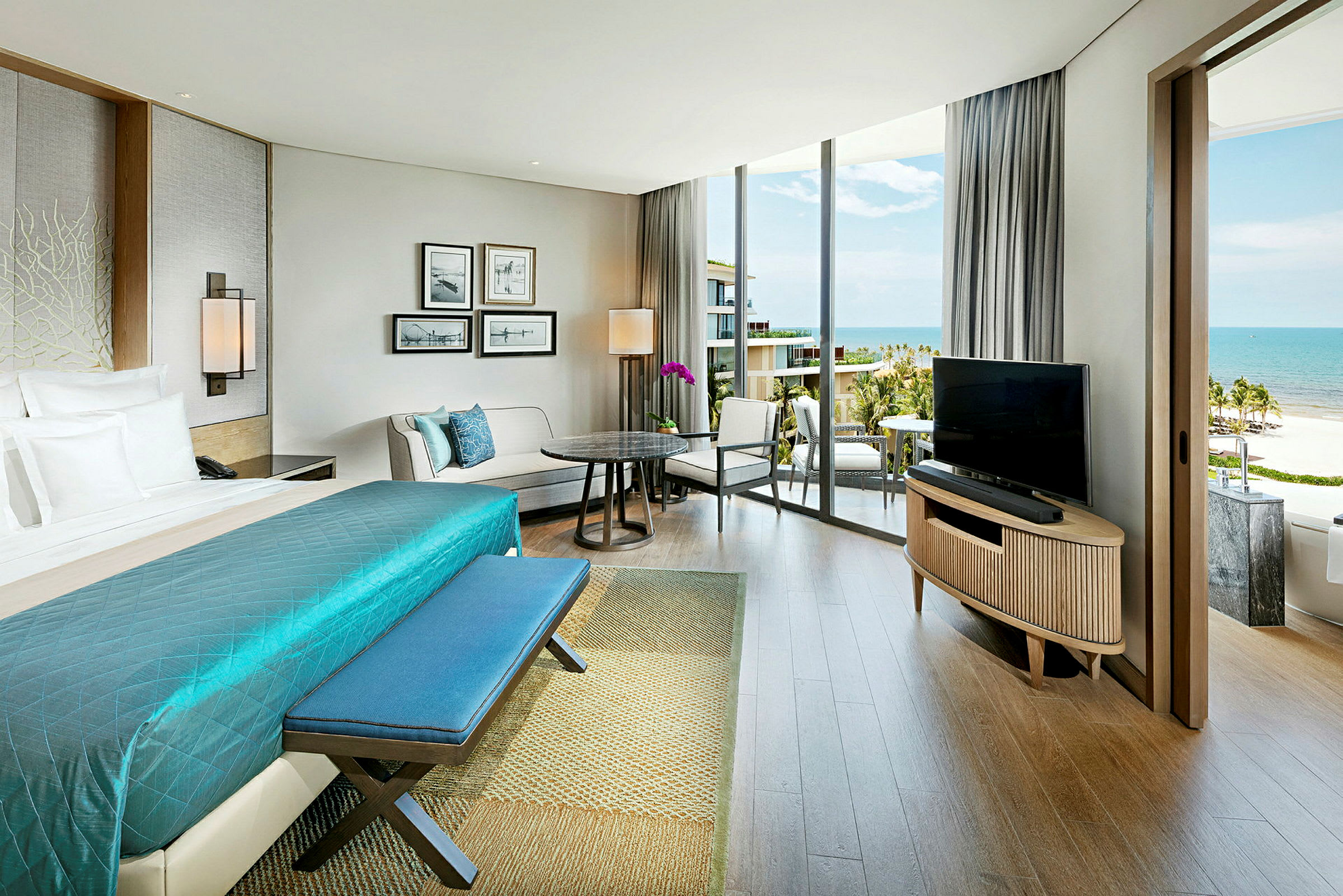 Ett rymligt hotellrum med en stor säng med blå överkast, sittgrupp och skrivbord. Stora fönster som vetter mot balkongen med utsikt över havet. (AI-genererad text)