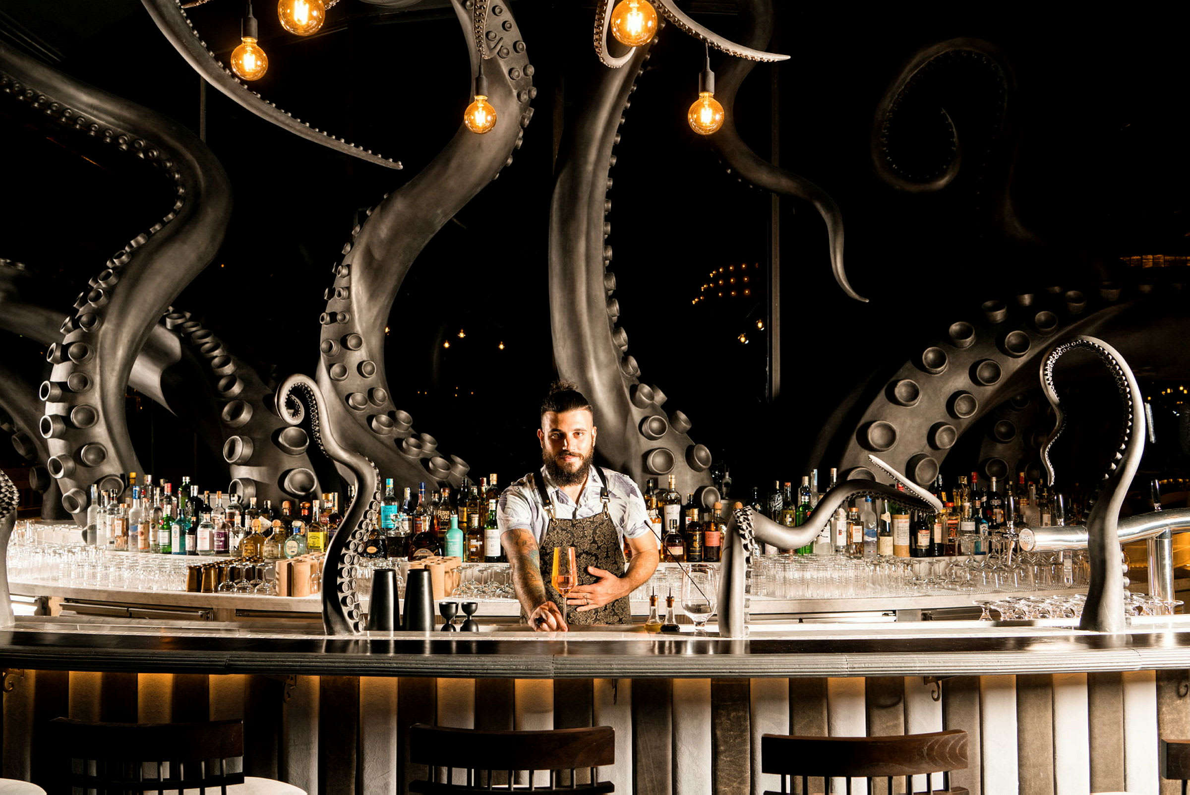 En bartender står vid en bar med detaljer av bläckfisktentakler, flaskor och glas syns bakom honom. (AI-genererad text)