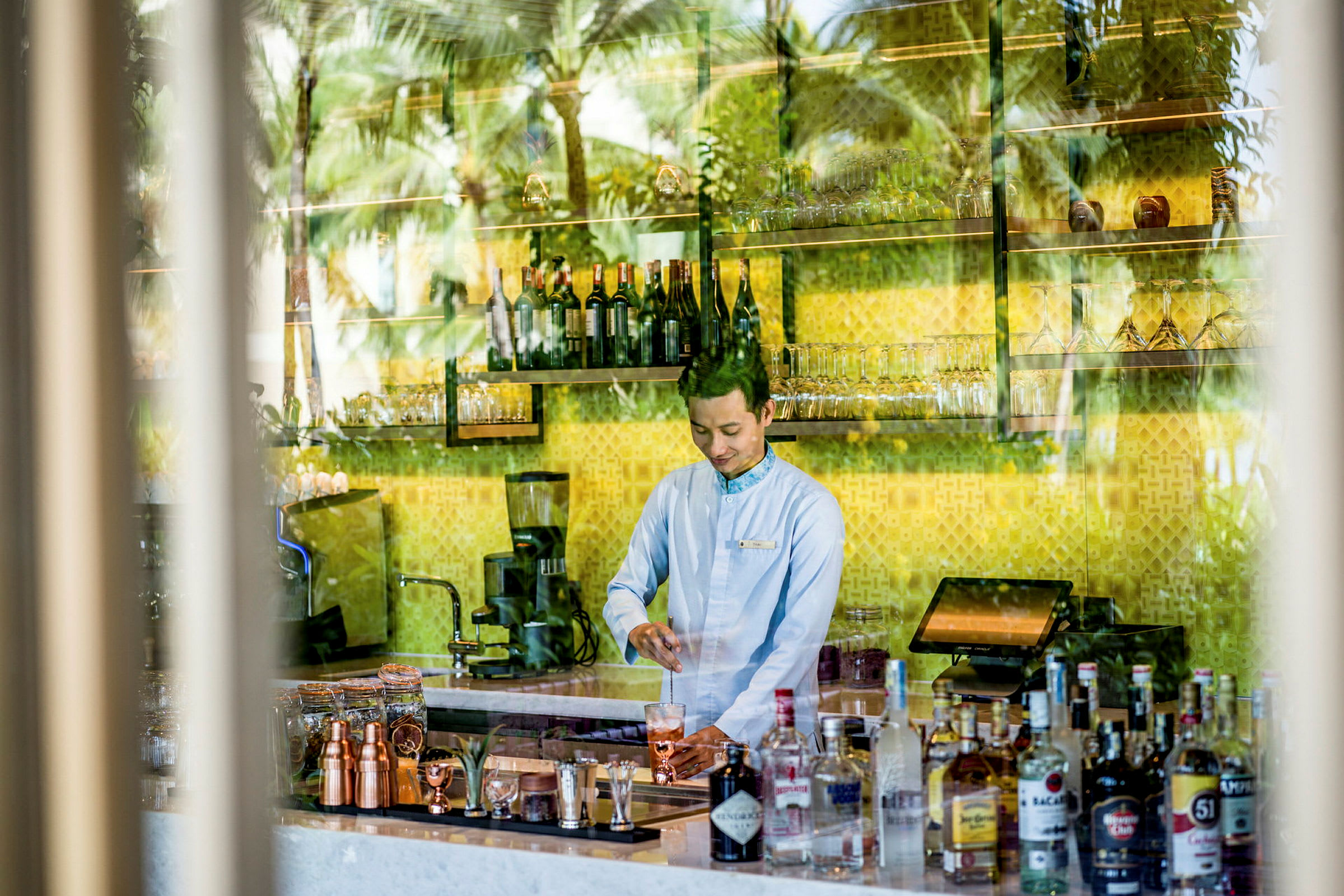 En bartender står bakom en bar fylld med flaskor, glas och tillbehör med en gul bakgrund. (AI-genererad text)
