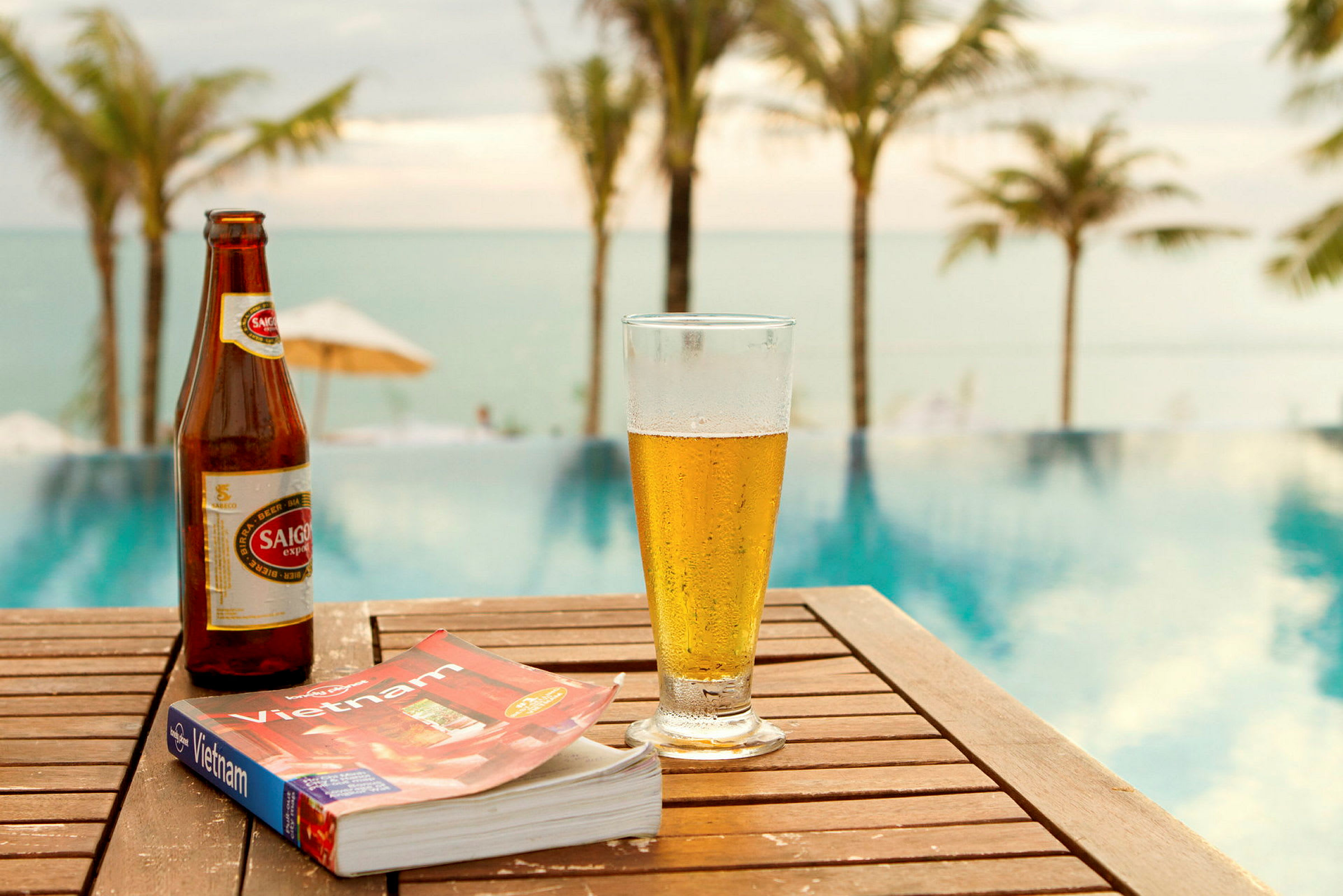 En flaska öl, ett glas öl och en bok med titeln "Vietnam" står på ett träbord vid en pool. I bakgrunden skymtas palmer och hav. (AI-genererad text)