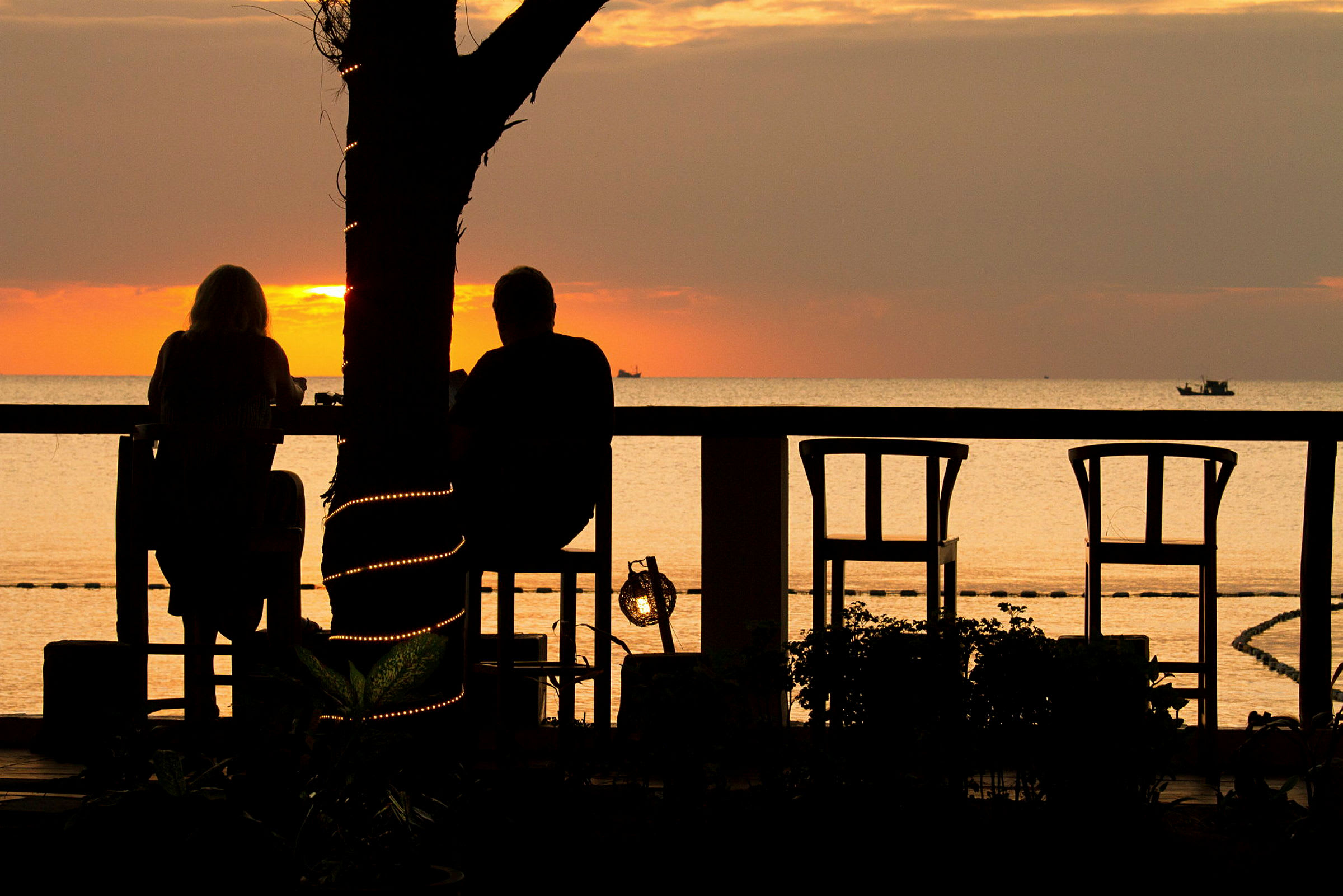 Två personer sitter vid ett bord och njuter av en solnedgång vid havet. Ett träd med dekorativa ljus står i förgrunden. (AI-genererad text)