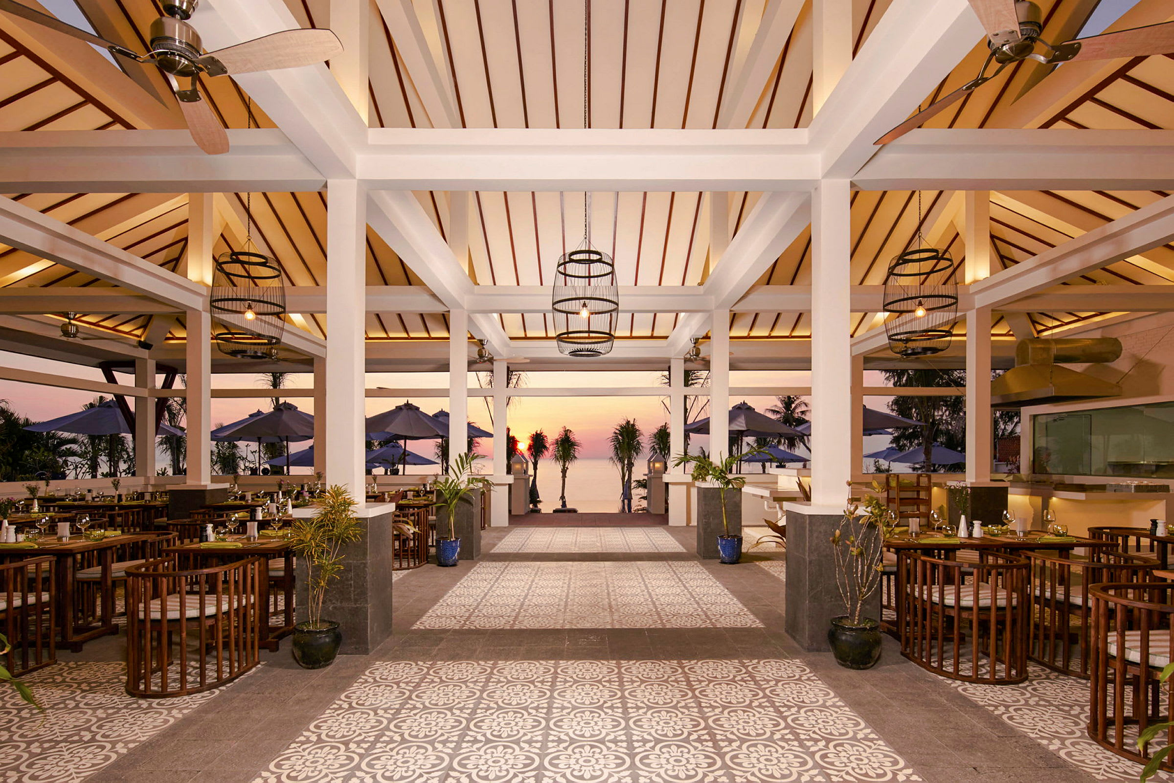 En restaurang med tak i trä, öppna väggar och golv med dekorativa mönster. Utanför syns bord, stolar, och parasoller samt solnedgång vid havet. (AI-genererad text)