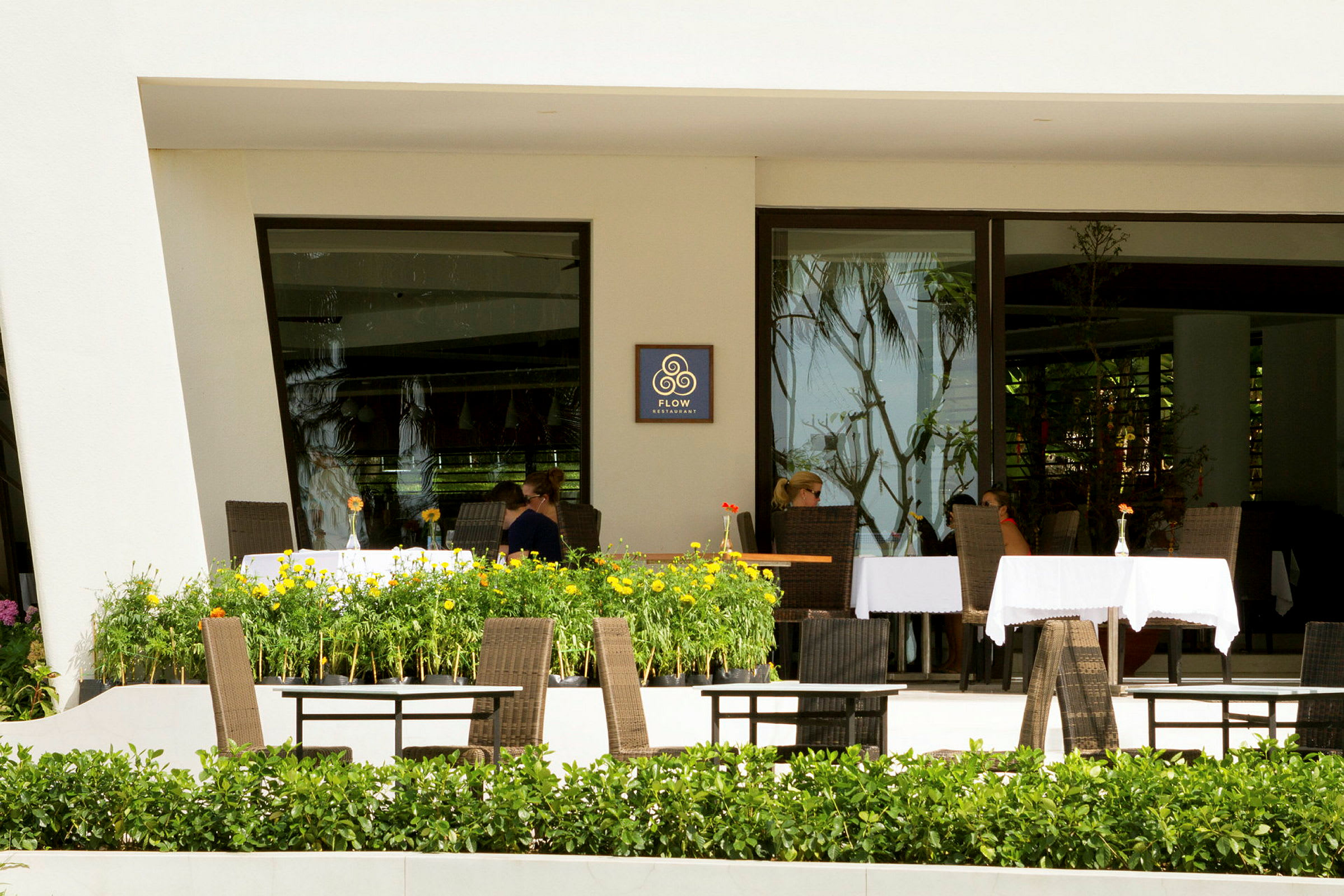 En restaurang med bord och stolar på en terrass omgiven av gröna växter och blommor. (AI-genererad text)