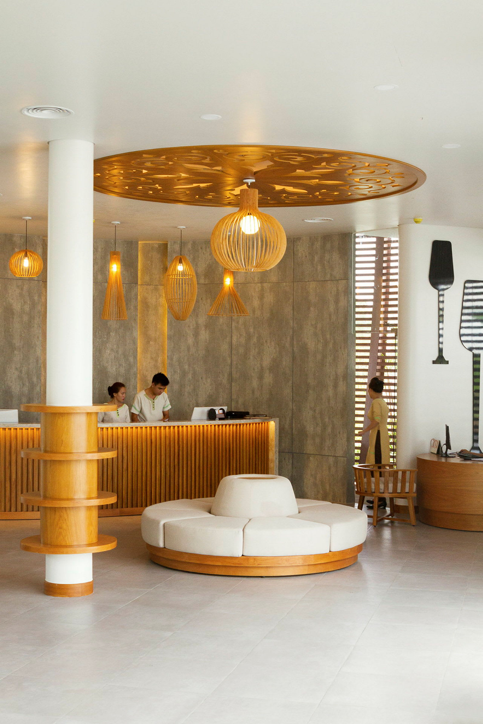 En hotellreception med en modern och stilren inredning. Det finns en bänk med personal, ett runt sittmöbel i mitten av rummet och dekorativa lampor i taket. (AI-genererad text)