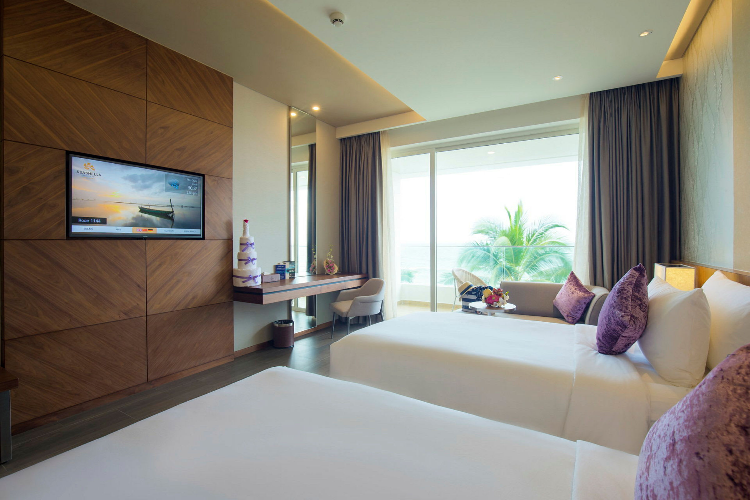 Hotellrum med två sängar, lila kuddar, skrivbord, stort fönster och utsikt över havet. (AI-genererad text)