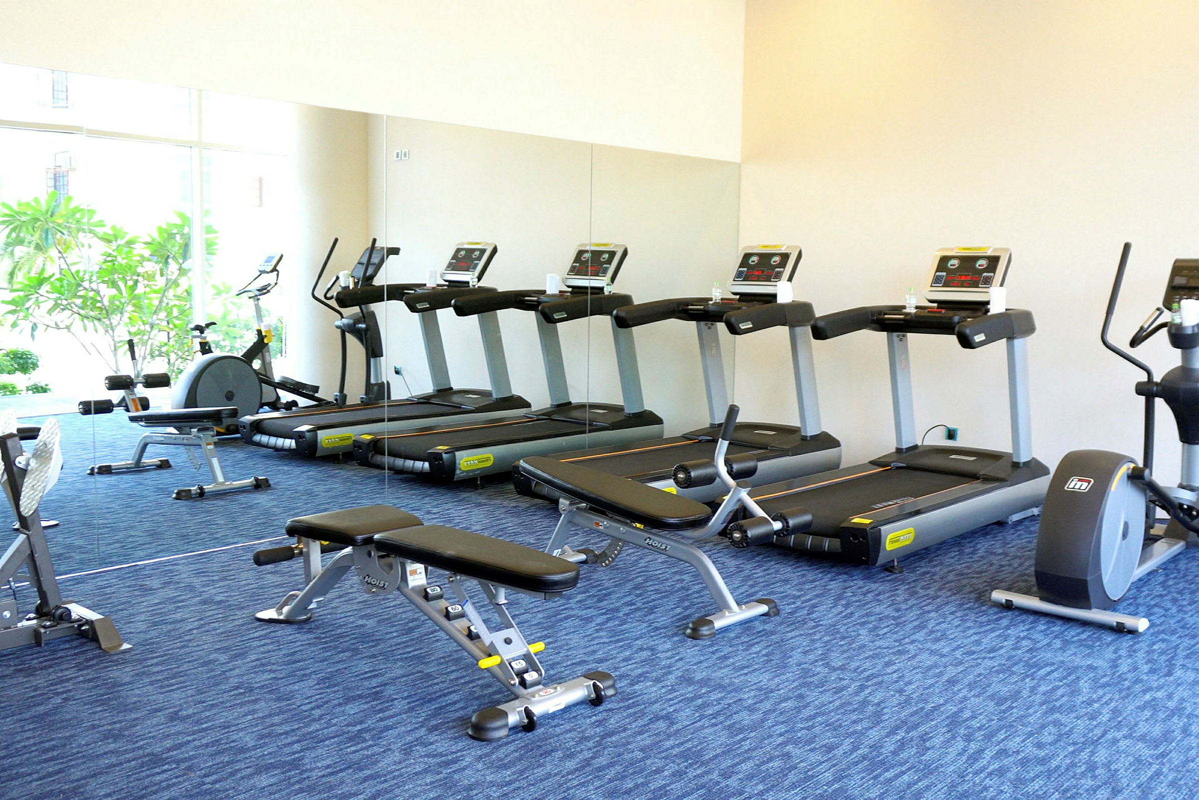 Ett gym med träningsutrustning som löpband och träningsbänkar, med spegelvägg och ljust fönster. (AI-genererad text)