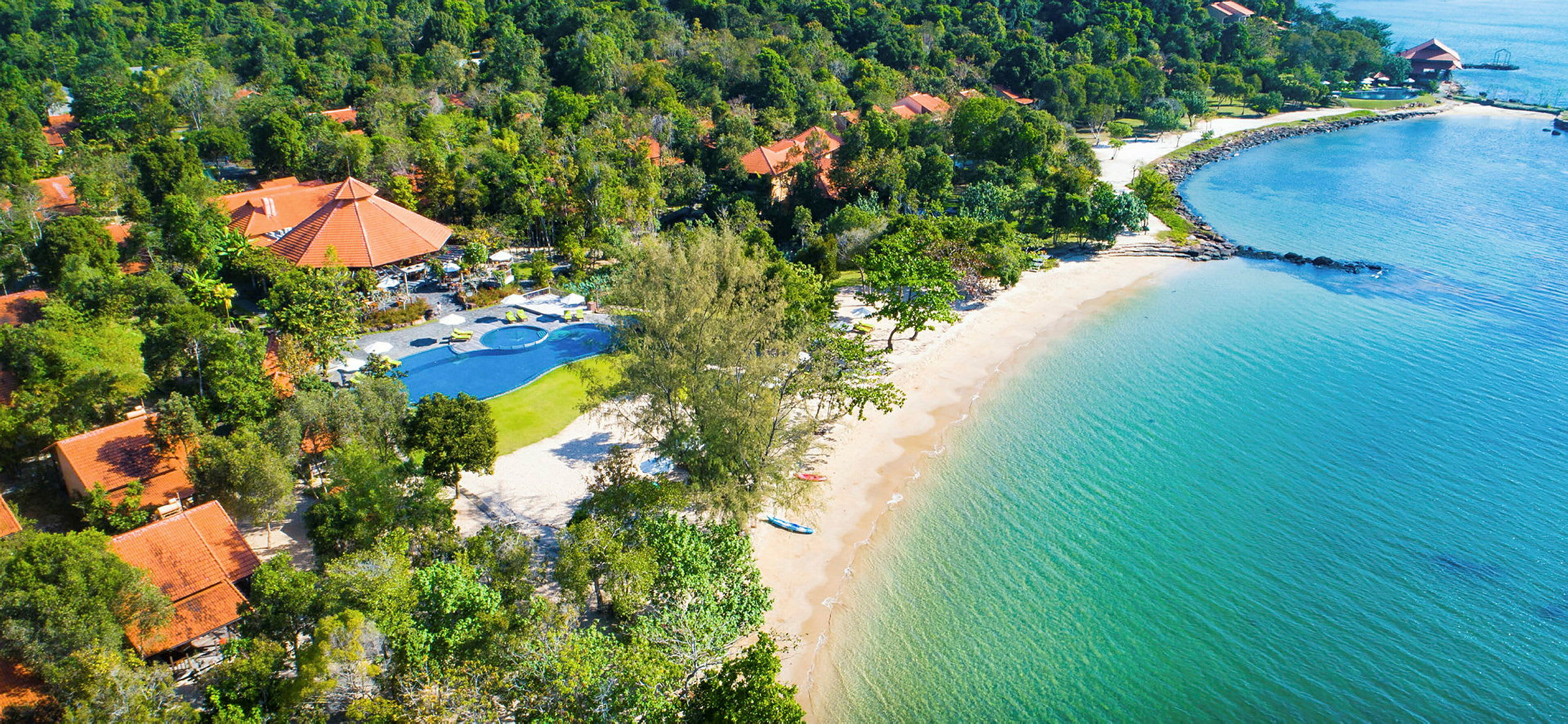 Flygfoto av strand med klart vatten, omgiven av grönskande träd och byggnader med röda tak. (AI-genererad text)