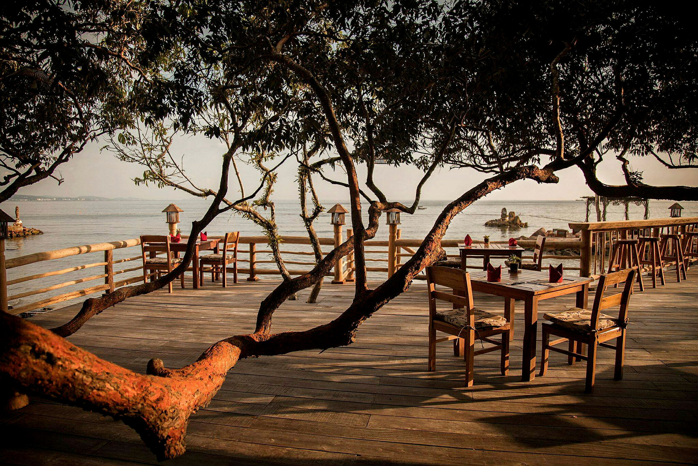 Ett trädäck med bord och stolar, omgiven av trädgrenar och med utsikt över havet. (AI-genererad text)