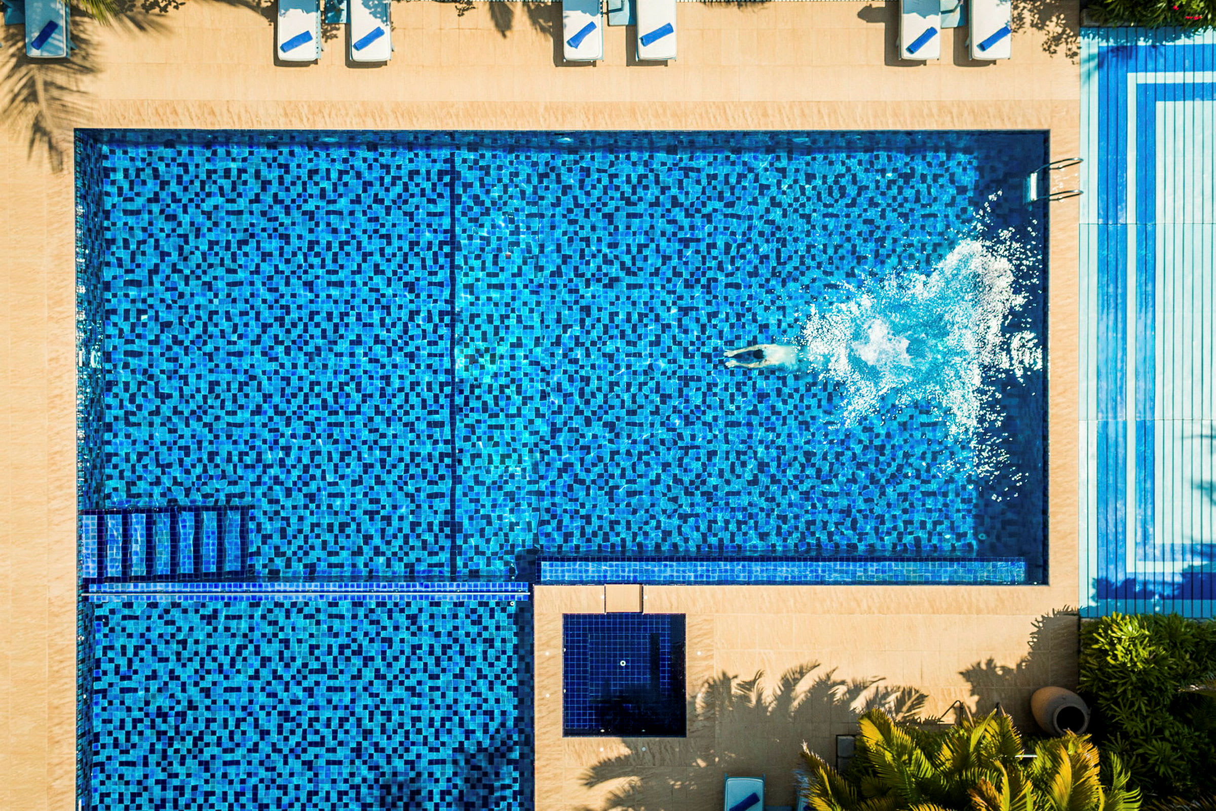 Flygfoto av en swimmingpool med blå mosaik och en person simmar i vattnet. (AI-genererad text)