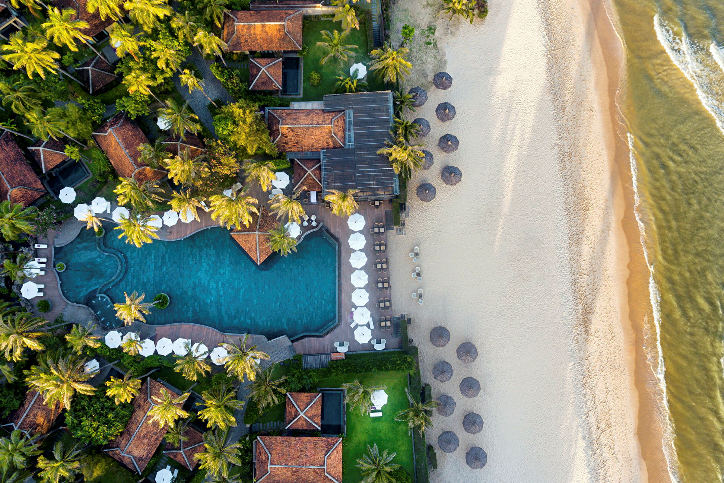 Flygfotografi av ett hotellområde med en pool, solstolar med parasoller och en sandstrand vid havet. (AI-genererad text)