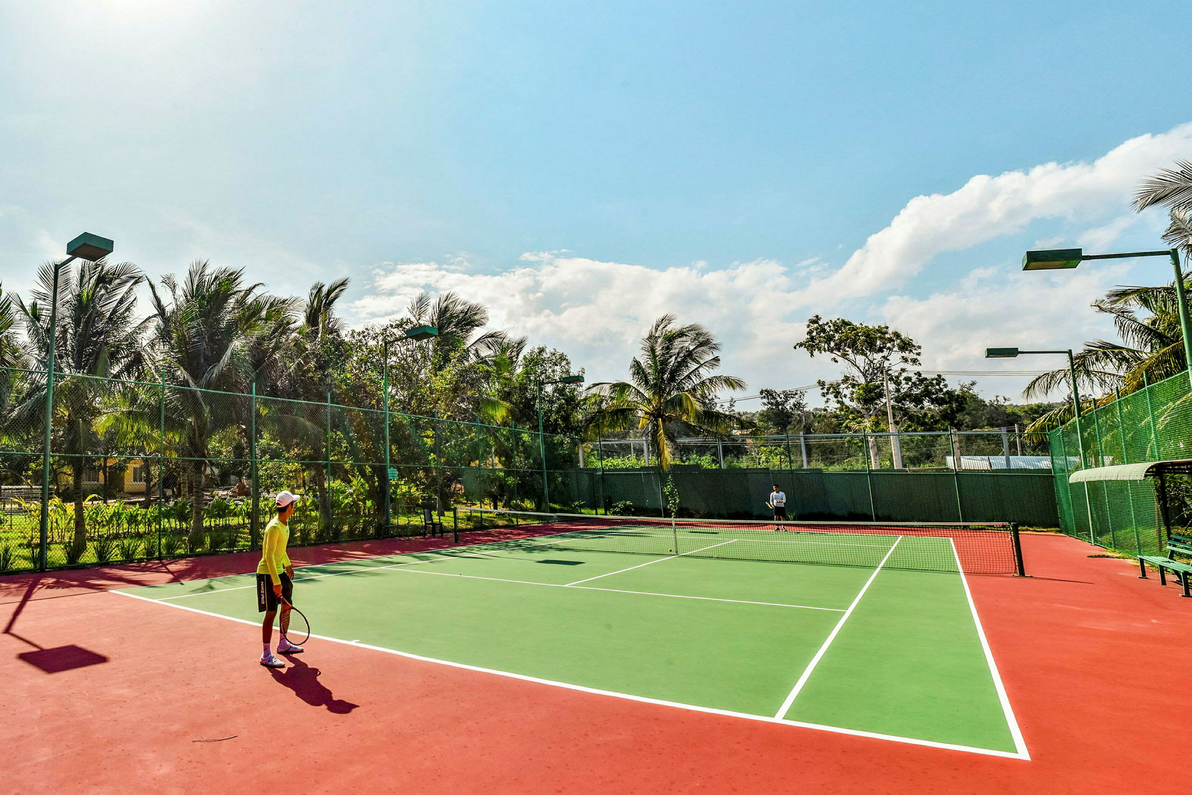 Två personer spelar tennis på en utomhusbana omgiven av grönska och palmer under en klar himmel. (AI-genererad text)