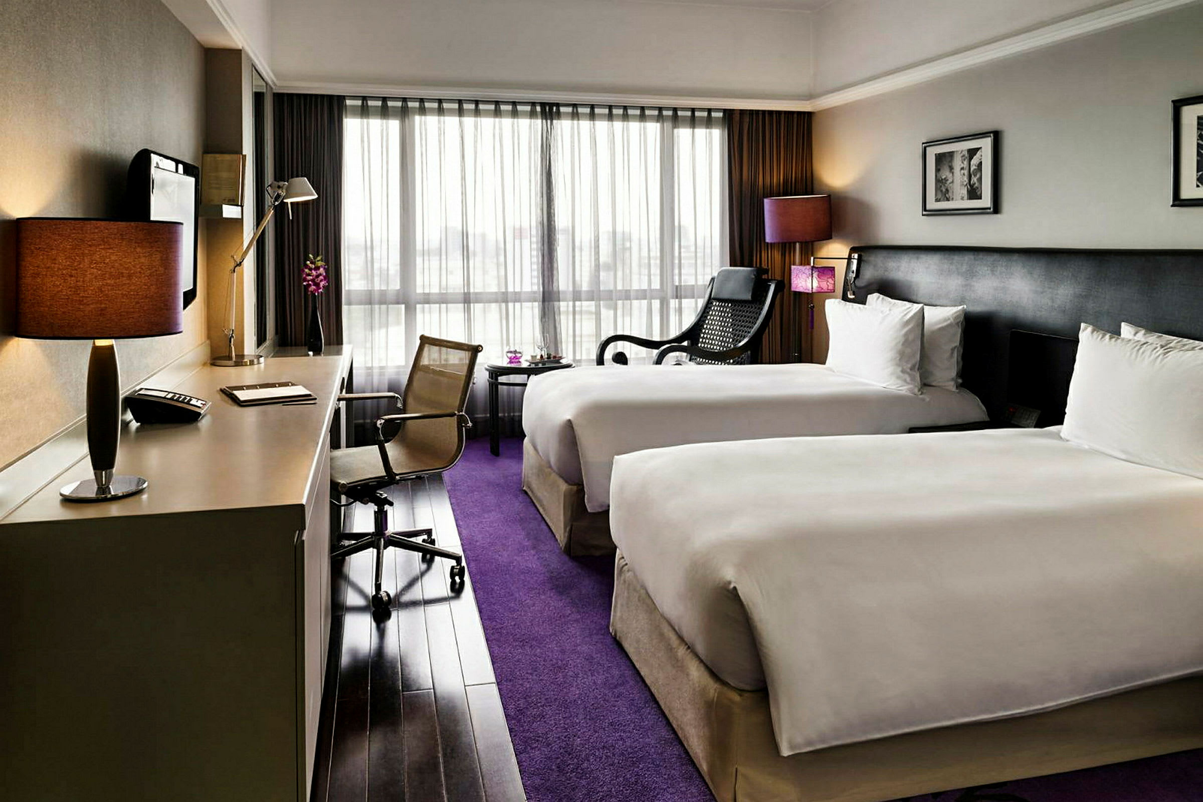 Ett hotellrum med två enkelsängar, ett skrivbord med stol, och en sittplats med fåtölj vid ett fönster. (AI-genererad text)