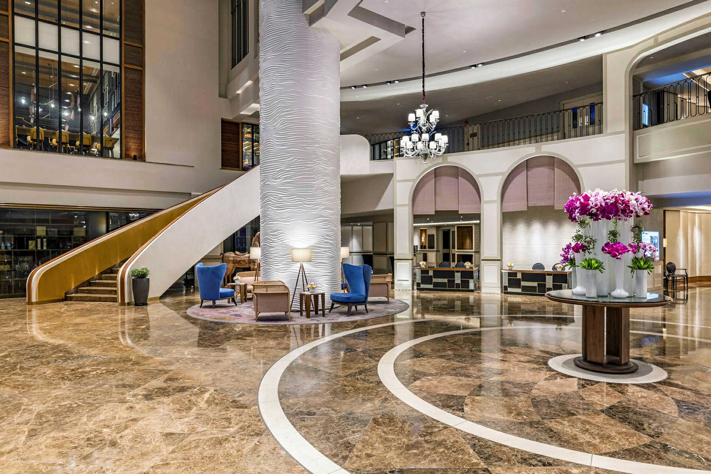 En hotellobby med marmorgolv, en stor pelare, en trappa, sittmöbler och en rund bordsdekoration med blommor. (AI-genererad text)